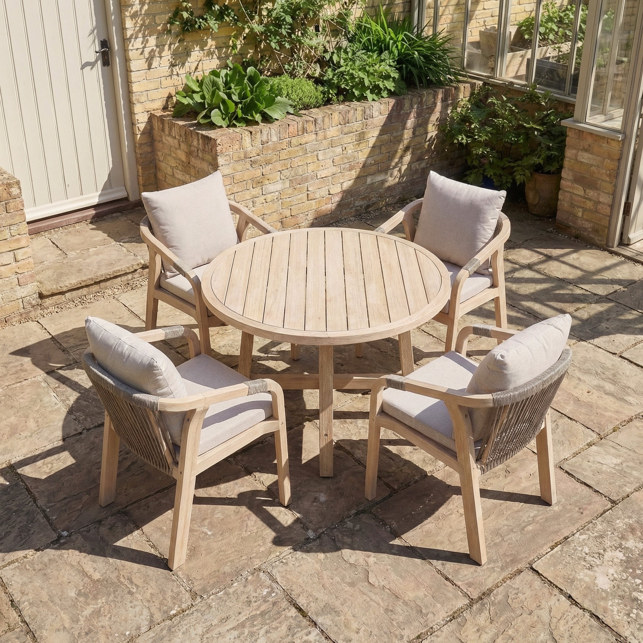 Quay 4 Seat Round Dining Set in Mocha: Features a round wooden table with four cushioned chairs, ideal for outdoor gatherings on your patio surrounded by greenery and brick walls, creating a warm and inviting dining space.