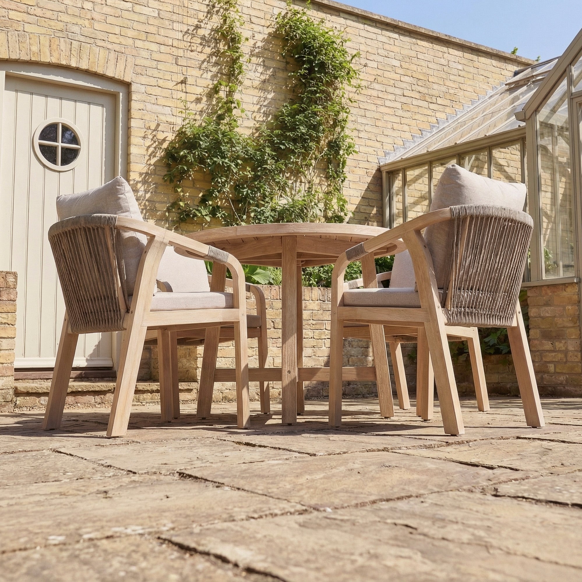 Outdoor patio with a Quay 4 Seat Round Dining Set in Mocha, featuring cushioned wooden chairs and a round table on stone flooring beside a charming brick house.