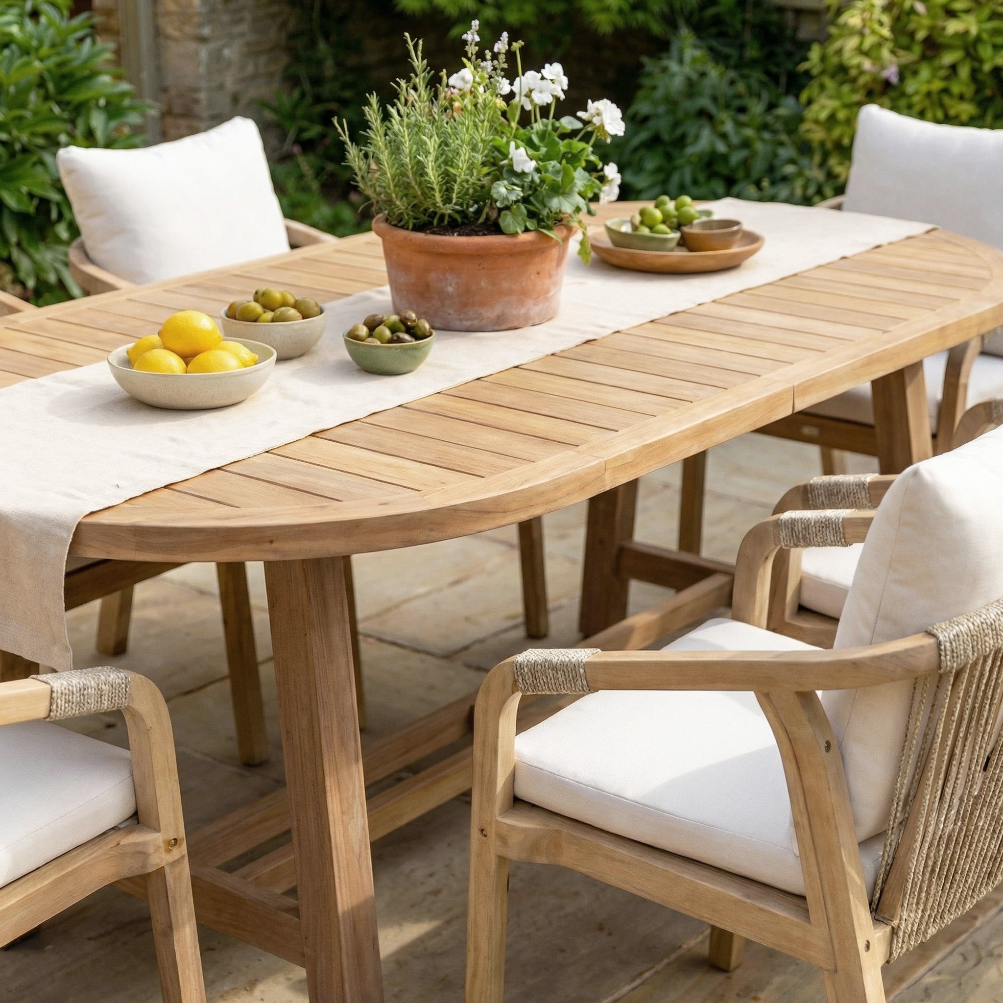Gather around the Quay 6 Seat Oval Dining Set in Linen, an elegant outdoor set featuring white cushions and a fresh tablescape with a potted plant plus bowls of lemons and olives.