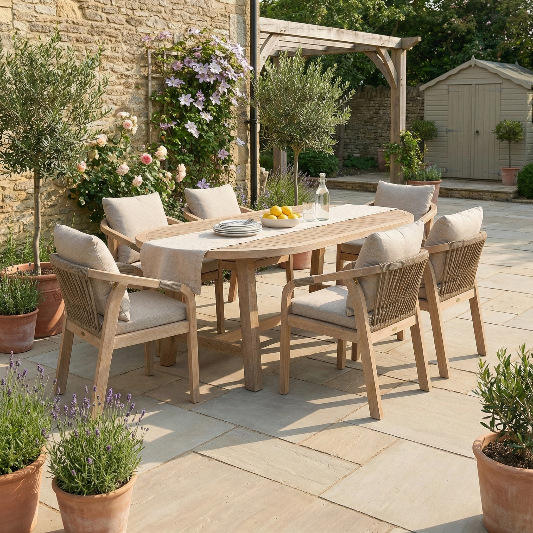 The Quay 6 Seat Oval Dining Set in Mocha, featuring six chairs, is arranged on a stone patio amid potted plants and greenery.