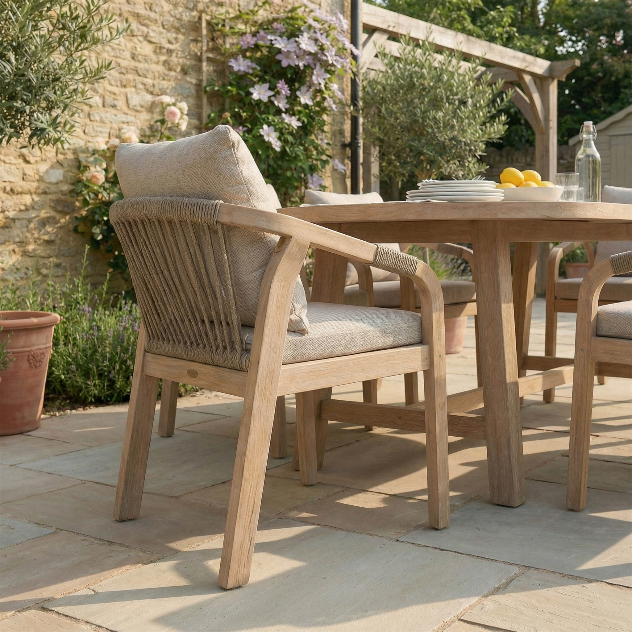 The Quay 6 Seat Oval Dining Set in Mocha features eucalyptus garden furniture with beige cushions, set on a stone patio beside lush plants—perfect for elevating any outdoor dining experience.