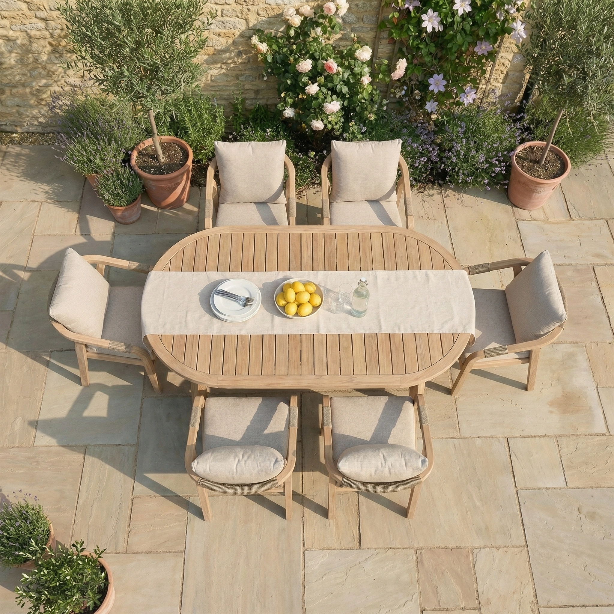 The Quay 6 Seat Oval Dining Set in Mocha features a wooden table and six cushioned chairs, ideal for outdoor relaxation on a stone patio. Lemons and a bottle add the perfect finishing touch.