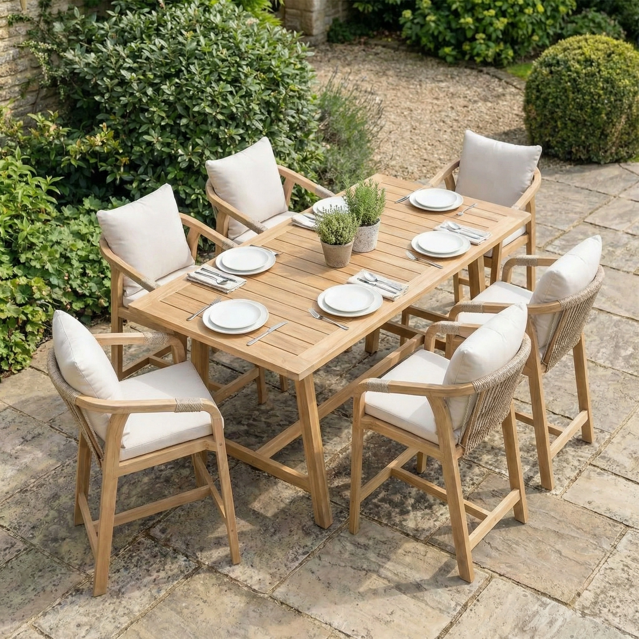 The Quay 6 Seat Rectangular Bar Set in Linen features an acacia wood table and six cushioned chairs, beautifully arranged with plates, cutlery, and potted plants on a stone patio.