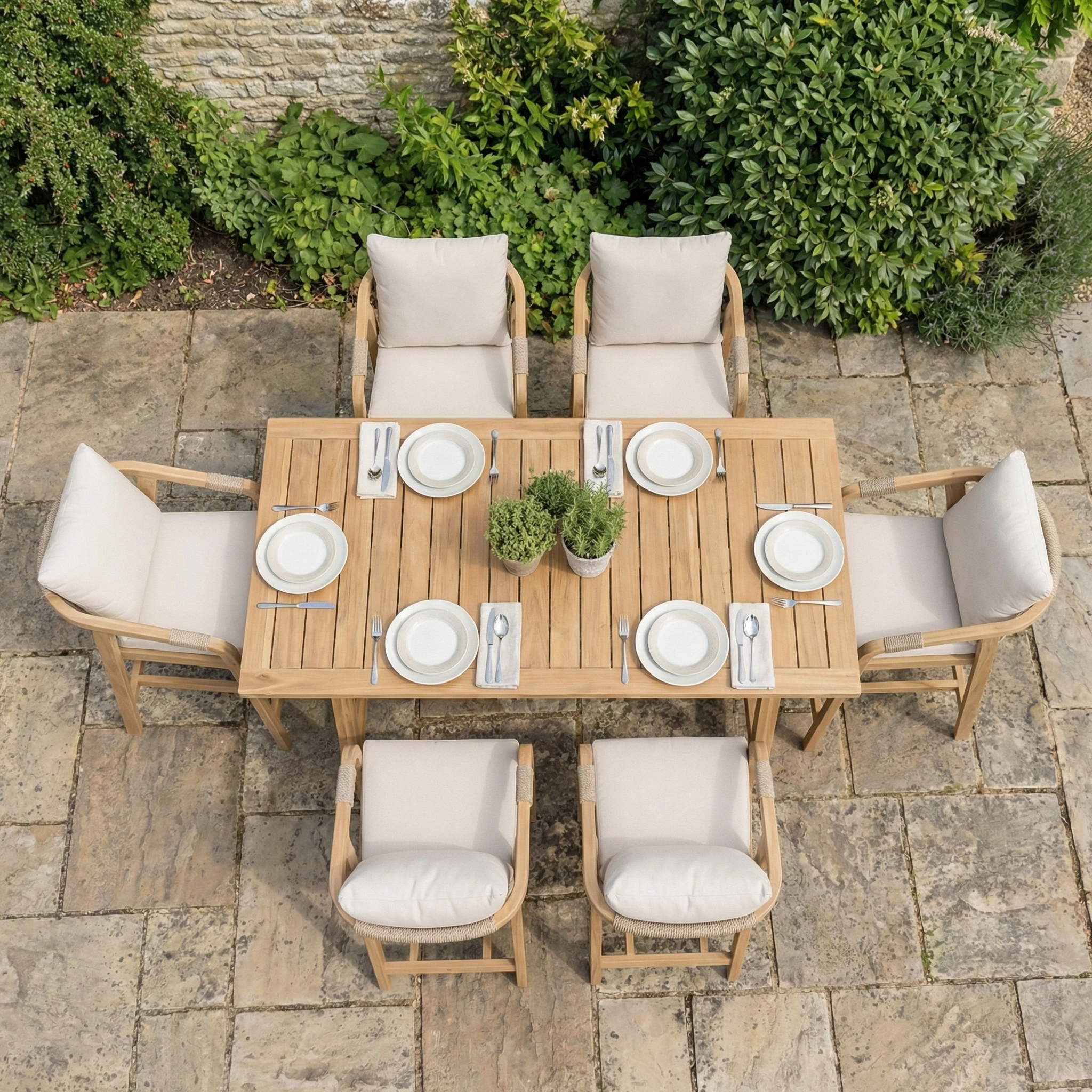 The Quay 6 Seat Rectangular Bar Set in Linen, styled with white plates, cutlery, and potted plants, sits elegantly on a stone patio.
