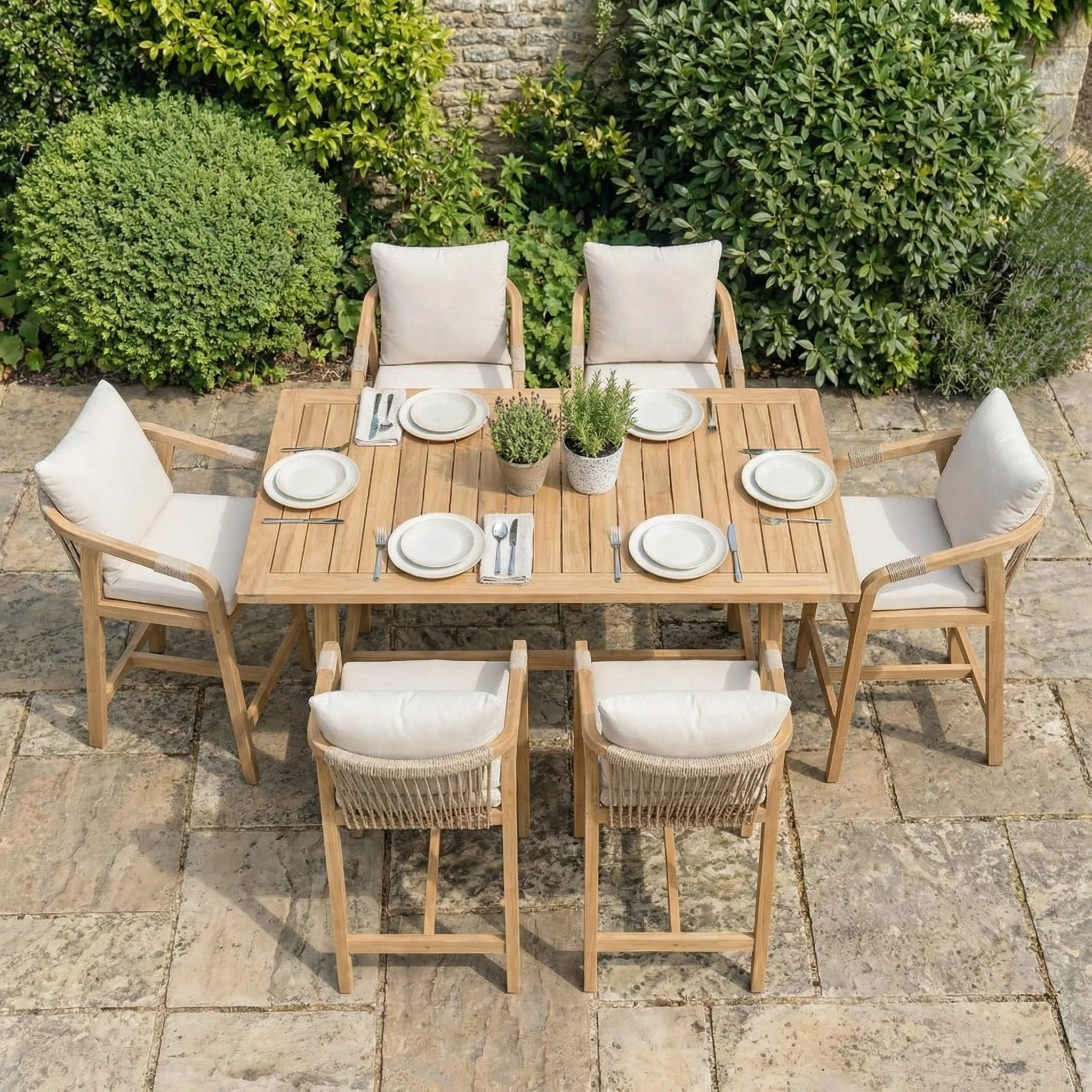The Quay 6 Seat Rectangular Bar Set in Linen, featuring cushioned chairs, is arranged on a stone patio alongside plates and potted plants.