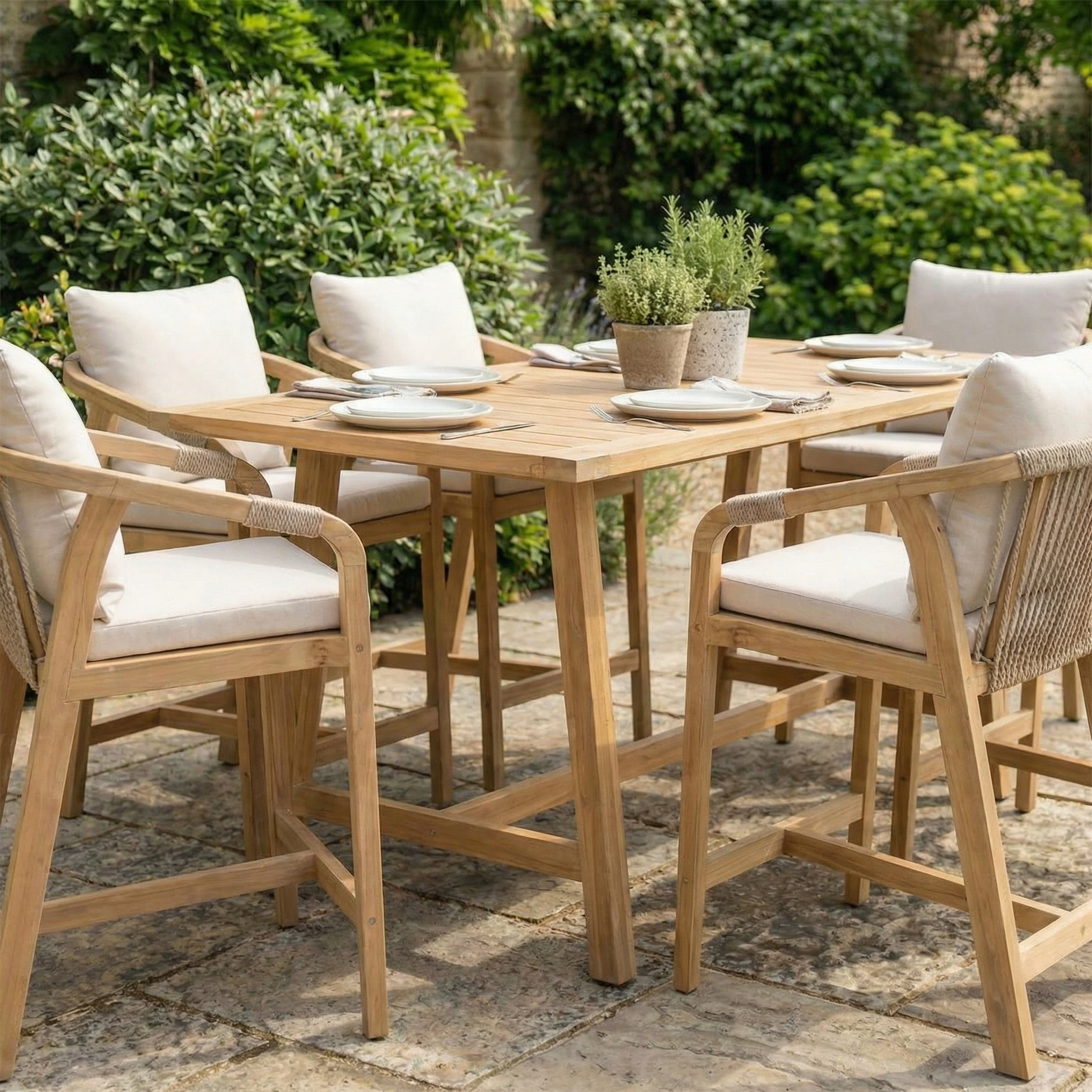 The Quay 6 Seat Rectangular Bar Set in Linen features six cushioned chairs, perfect for outdoor dining on a stone patio with potted plants and lush greenery.