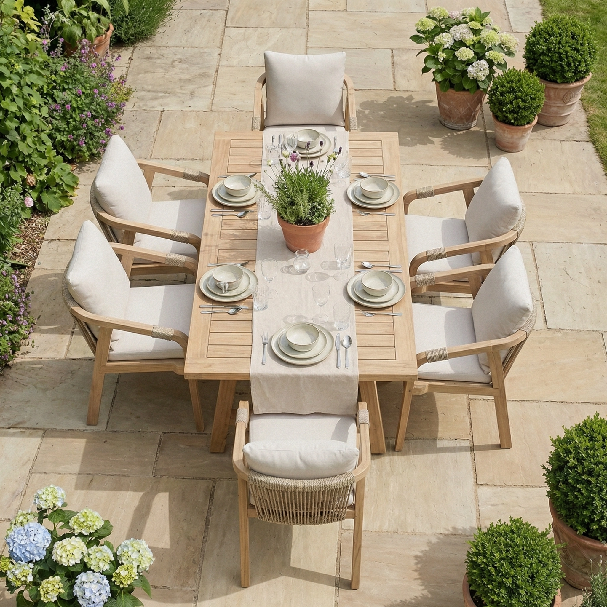 The Quay 6 Seat Rectangular Dining Set in Linen features a wooden table and six cushioned chairs made from durable acacia wood, perfect for outdoor dining among lush greenery and flowers.