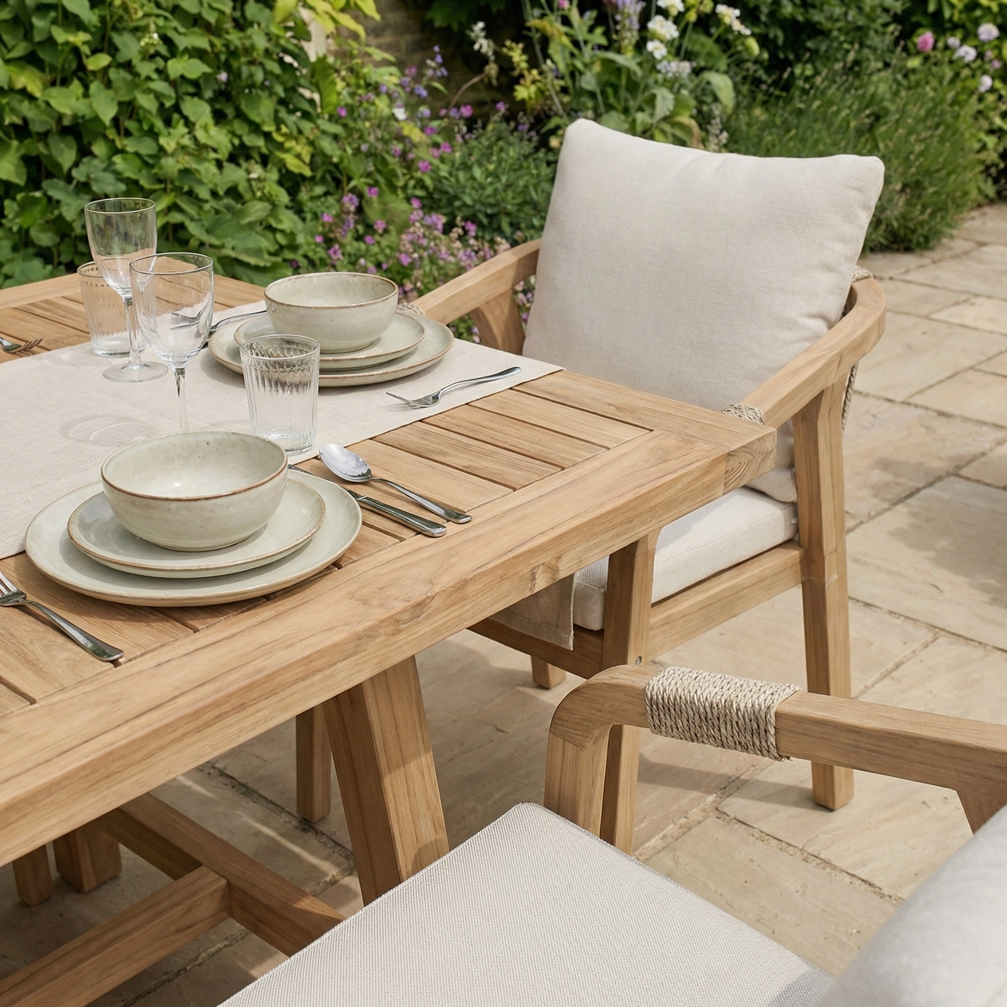 The Quay 6 Seat Rectangular Dining Set in Linen offers cushioned seating and a sturdy table, perfect for outdoor dining on a stone patio surrounded by greenery.