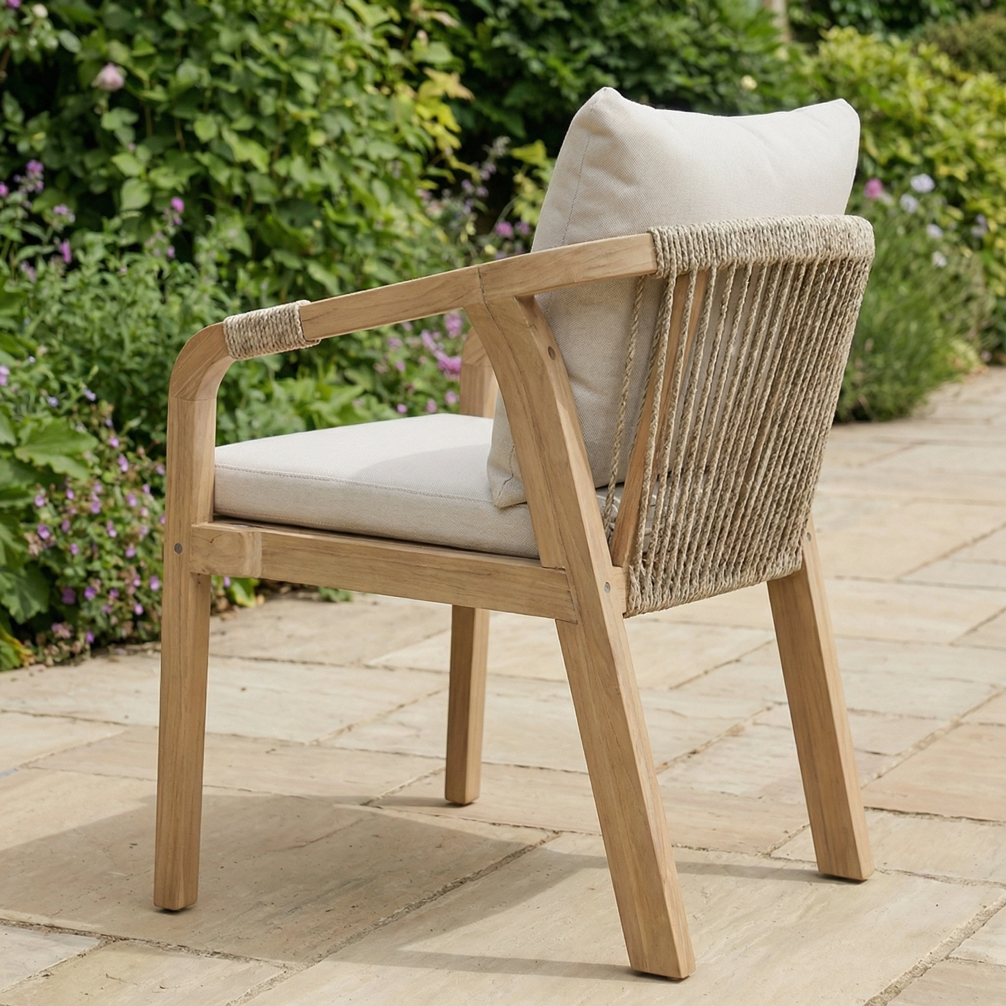 The Quay 6 Seat Rectangular Dining Set in Linen features acacia wood and beige cushions with rope detailing, ideal for outdoor dining on a stone patio surrounded by greenery.