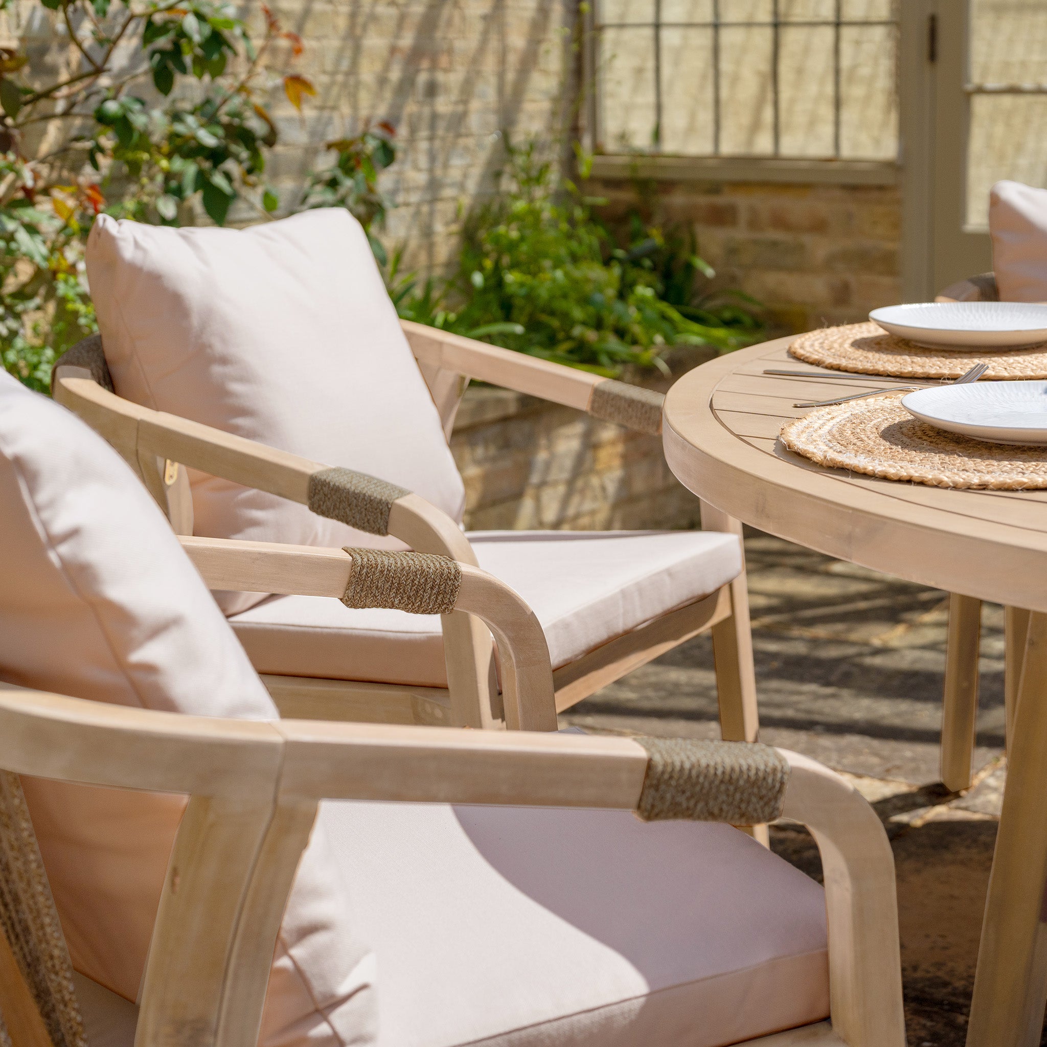 The Quay 6 Seat Round Dining Set in Mocha, arranged with plates and woven placemats, creates an inviting spot for outdoor dining in a sunny garden.
