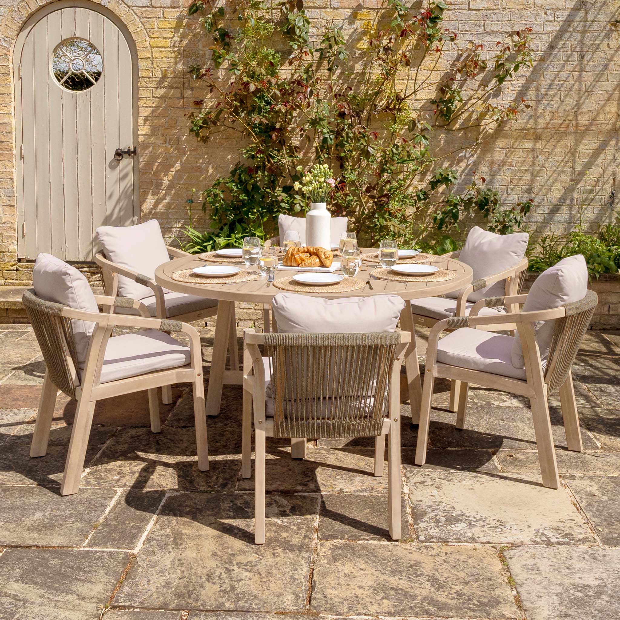 The Quay 6 Seat Round Dining Set in Mocha, with cushioned chairs, is ideal for memorable outdoor gatherings on a stone terrace by a vine-covered wall, offering an inviting and elegant al fresco dining spot.