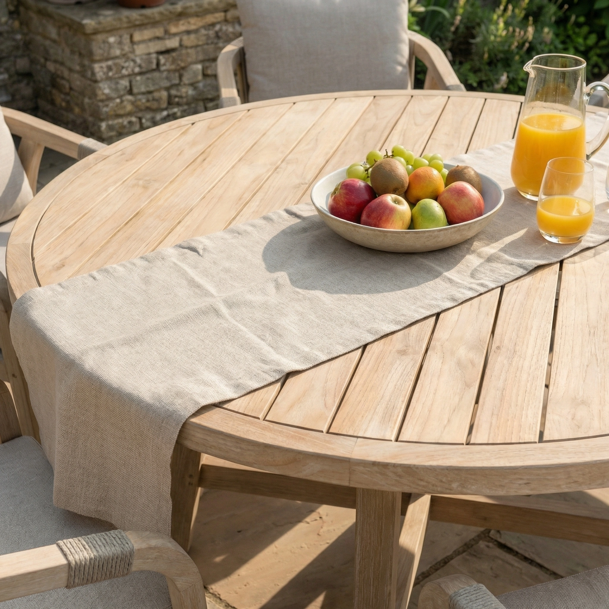 The Quay 6 Seat Round Dining Set in Mocha features a wooden table with a beige runner, set with a bowl of fruit, glass, and jug of orange juice, and is surrounded by stylish eucalyptus dining chairs.