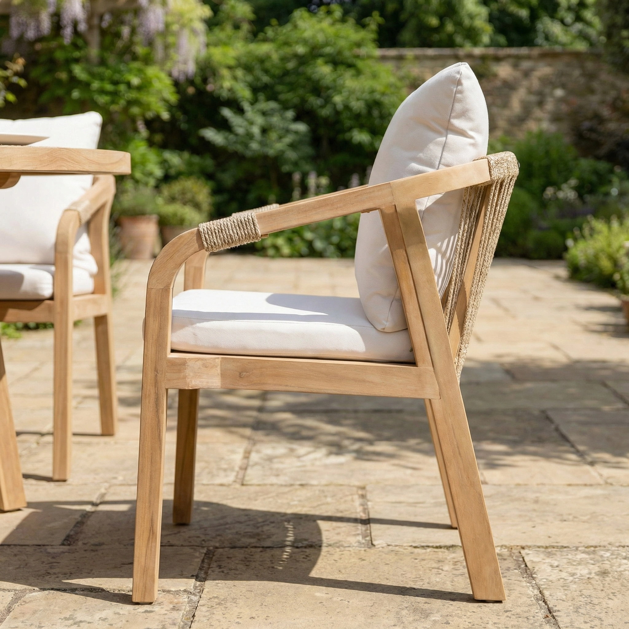 The Quay 6 Seat Round Dining Set in Linen features acacia wood chairs with white cushions, placed on a stone patio and surrounded by greenery and a brick wall for an inviting outdoor dining or relaxing space.