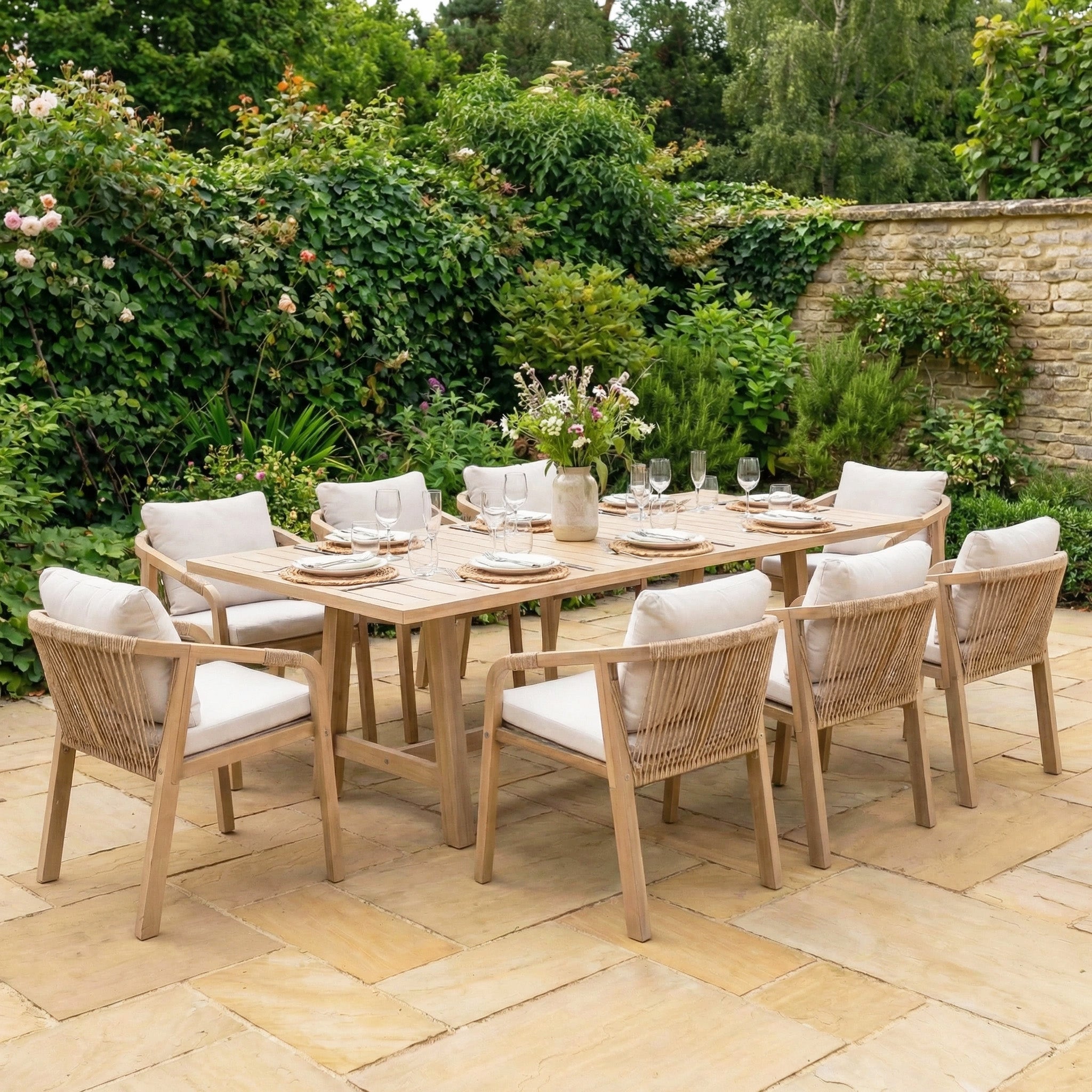 The Quay 8 Seat Rectangular Dining Set in Linen includes eight cushioned chairs and a spacious acacia wood table, perfect for outdoor gatherings on a stone patio amid lush greenery and flowers.