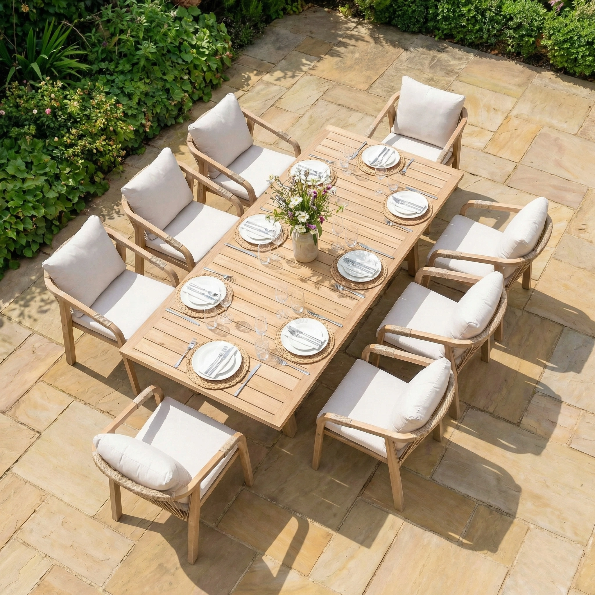 The Quay 8 Seat Rectangular Dining Set in Linen includes an acacia wood table and eight cushioned chairs, stylishly set on a stone patio with plates and glasses.