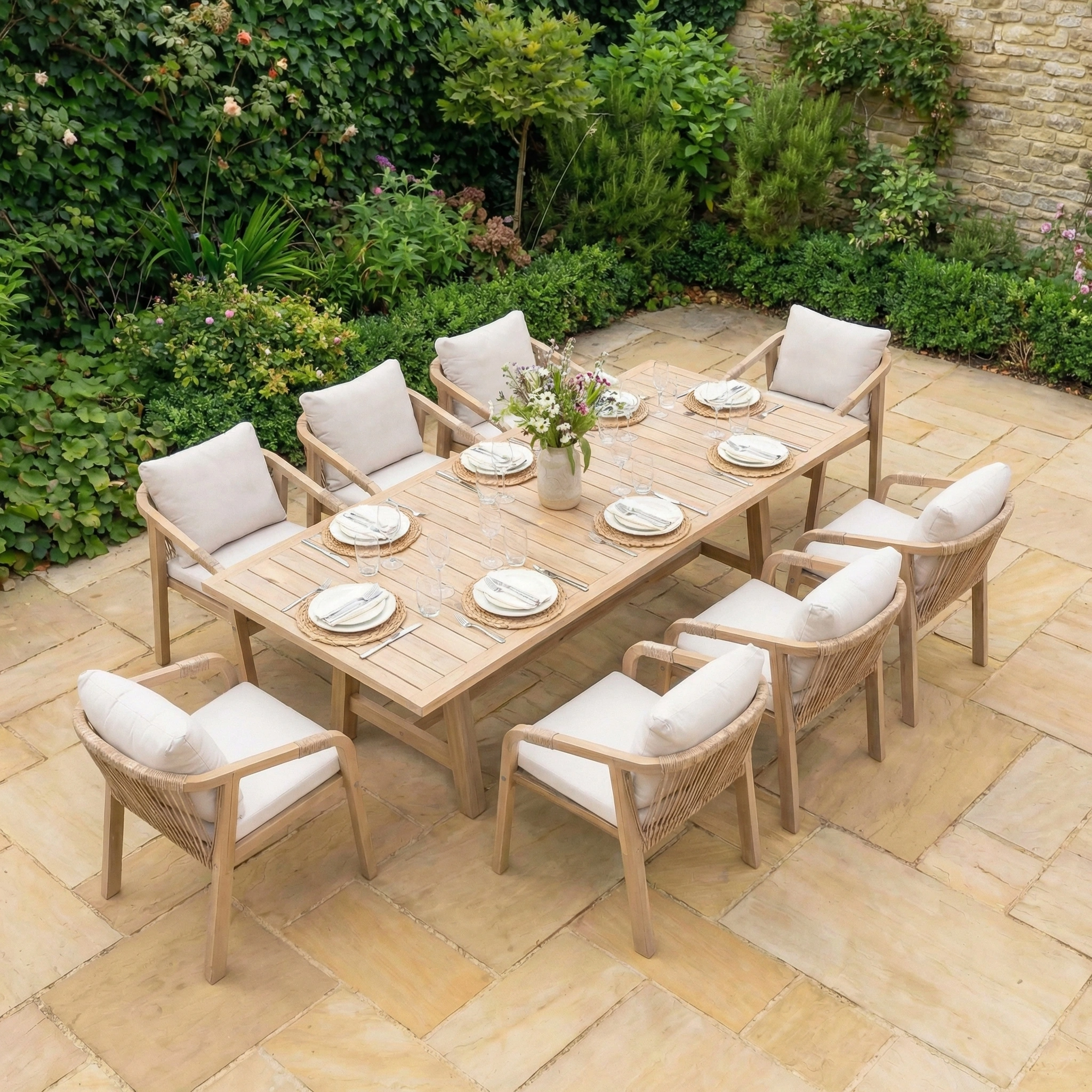 A Quay 8 Seat Rectangular Dining Set in Linen, featuring cushioned chairs, is arranged on a stone patio amidst lush greenery.