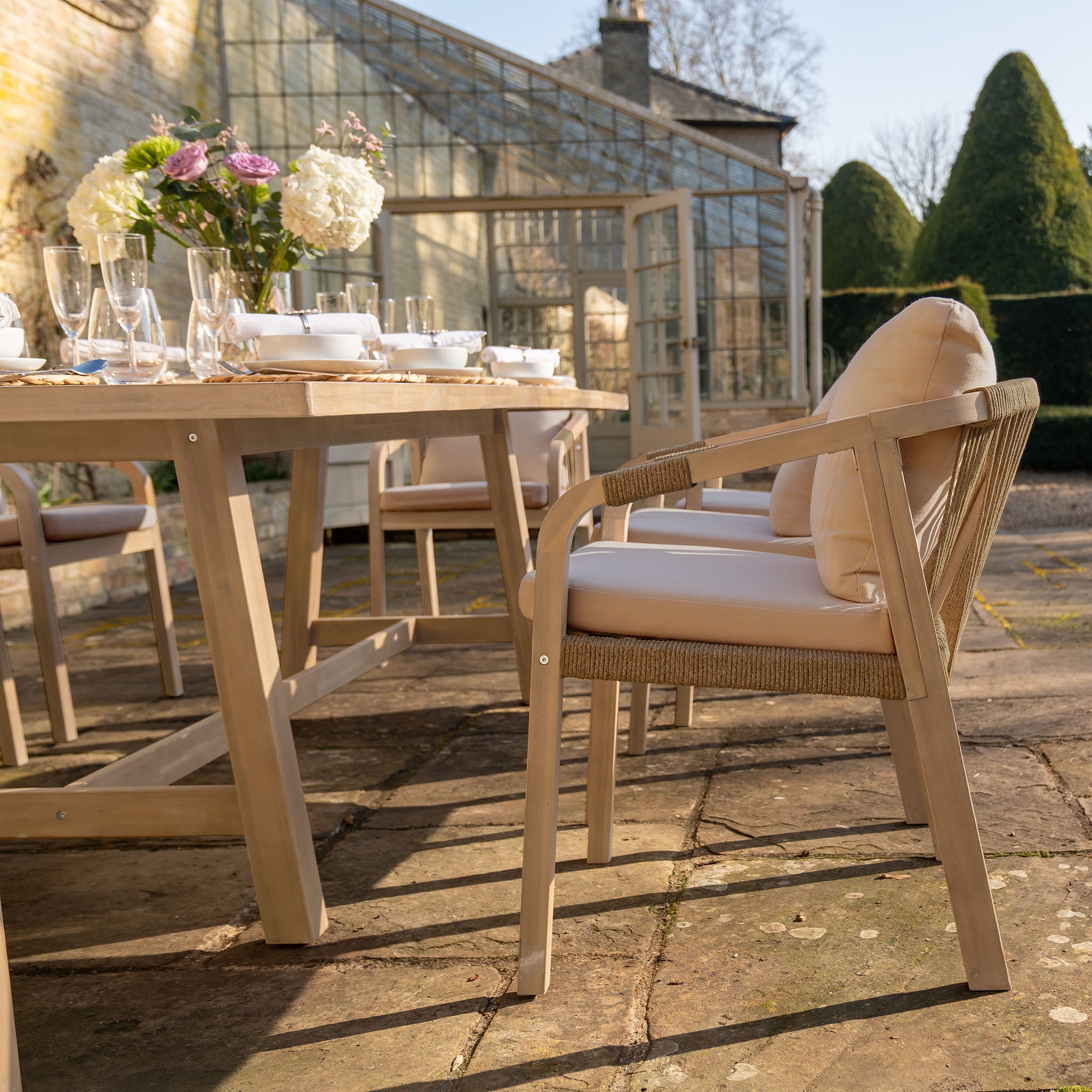 The Quay 8 Seat Rectangular Dining Set in Mocha, featuring cushioned chairs and a stylish table, is beautifully arranged on a stone patio next to a glass greenhouse.