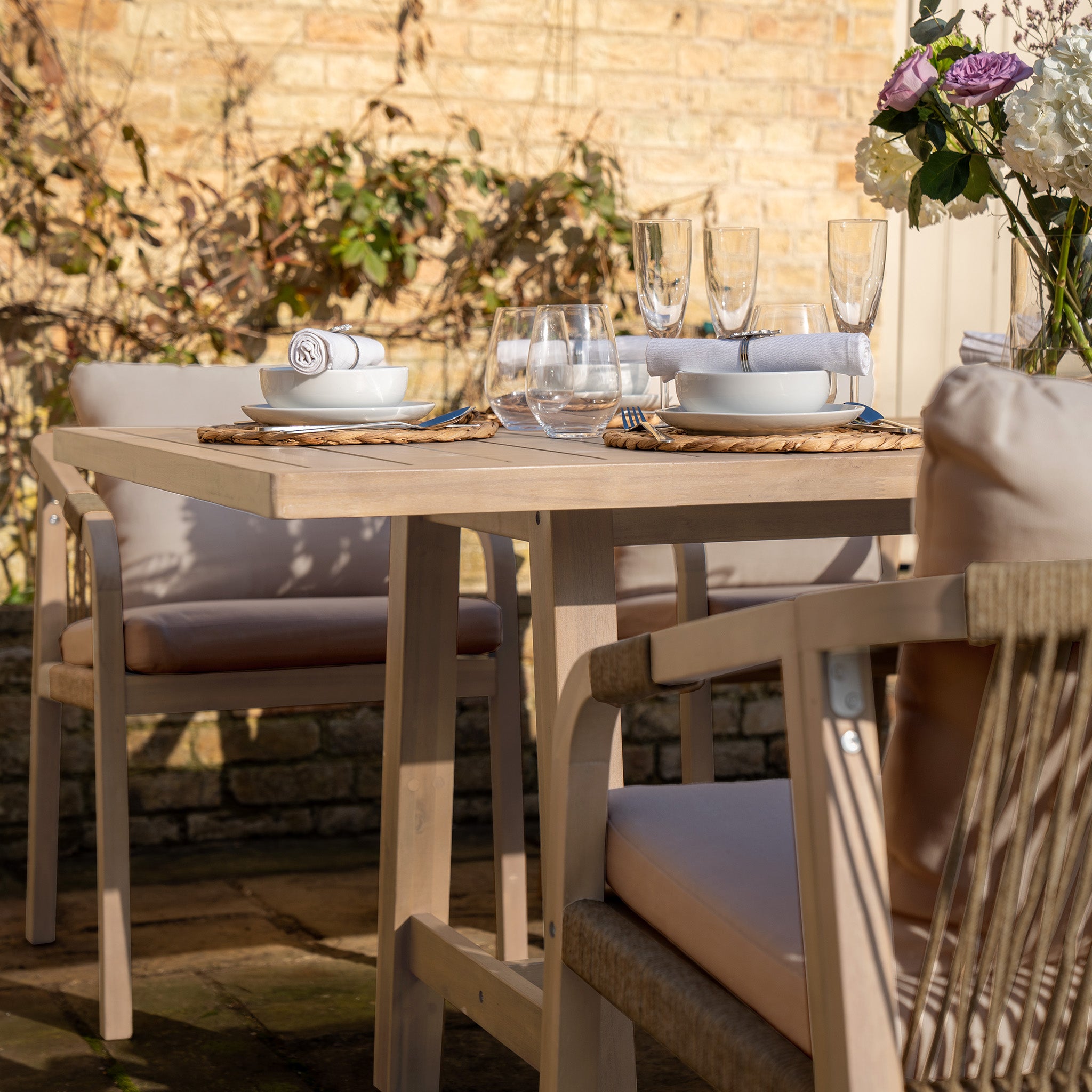 The Quay 8 Seat Rectangular Dining Set in Mocha features plates, glasses, and flowers on its elegant eucalyptus table, surrounded by cushioned chairs and bathed in sunlight.