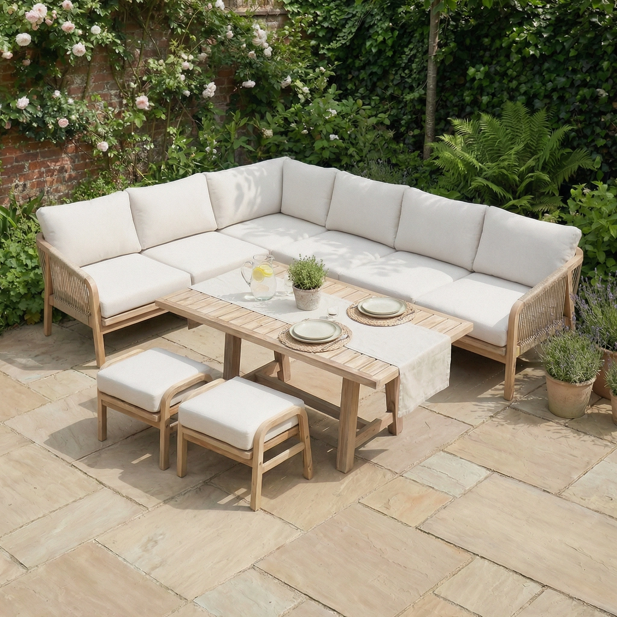 The Quay Corner Dining Set in Linen includes wooden patio furniture with white cushions, a table for two, and is set amid lush green garden plants.