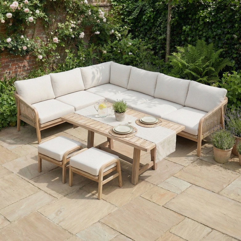 The Quay Corner Dining Set in Linen includes wooden patio furniture with white cushions, a table for two, and is set amid lush green garden plants.