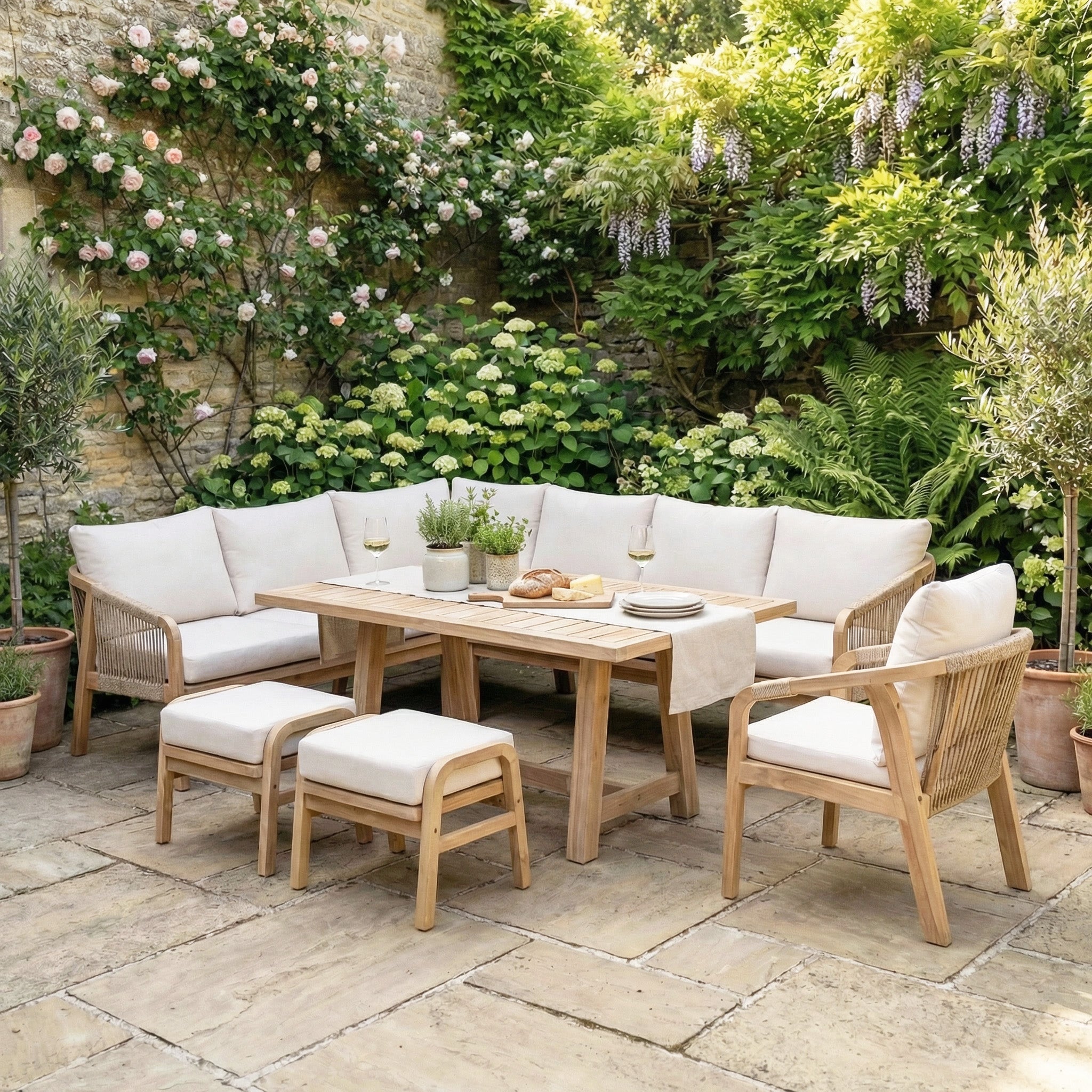 The Quay Corner Dining Set with Armchair in Linen creates a stylish corner seating area in a lush garden, enhanced by flowering plants and stone walls.