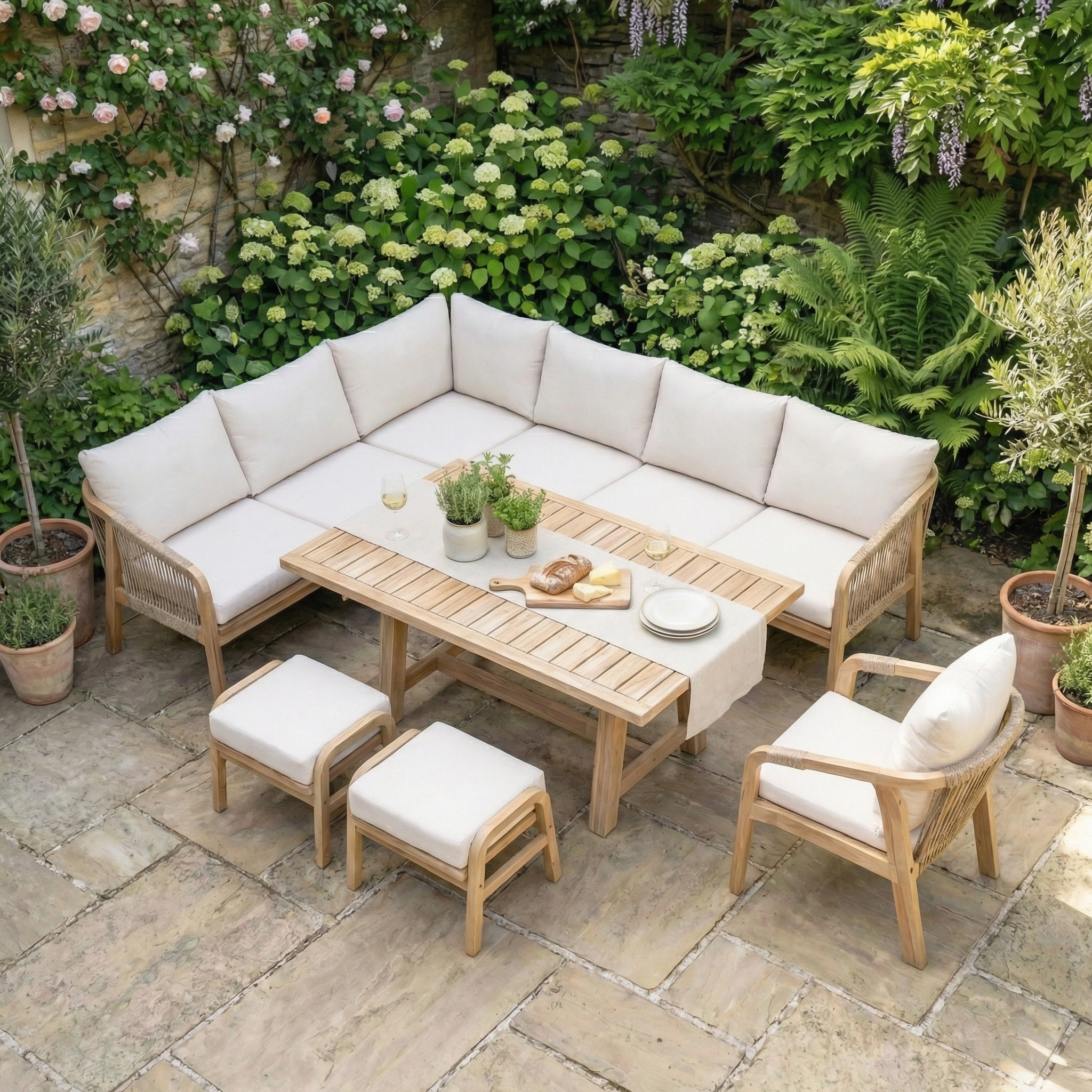 Outdoor patio featuring the Quay Corner Dining Set with Armchair in Linen, cushioned seating, an acacia wood table, potted plants, and lush greenery set in a stone-paved garden.