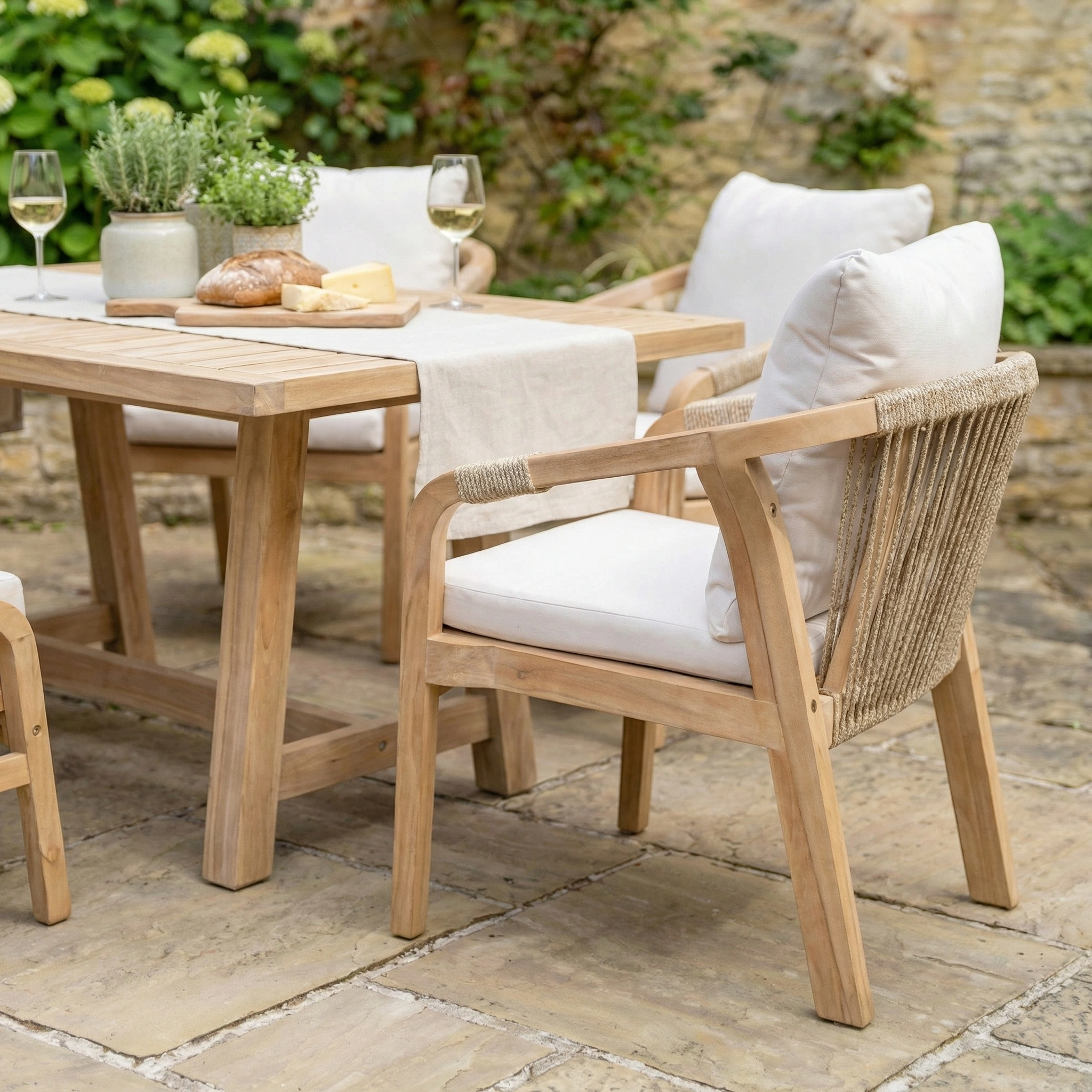 The Quay Corner Dining Set with Armchair in Linen is arranged on a stone patio, featuring cushioned chairs, wine glasses, bread, and cheese for relaxed outdoor dining.