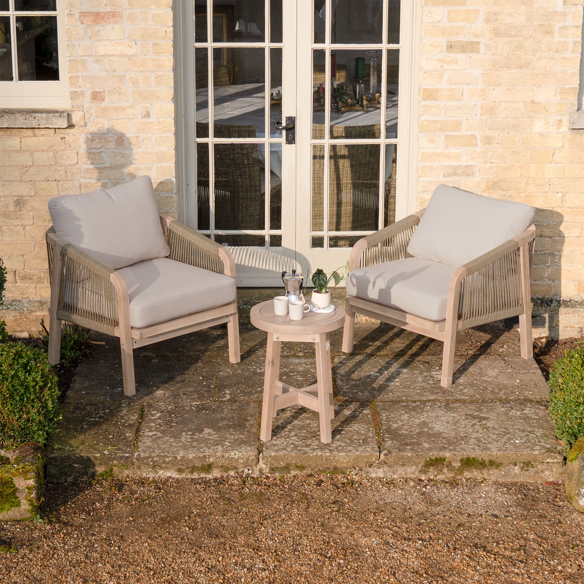 The Quay Lounge Set in Mocha includes two cushioned patio chairs and a small table with coffee cups, making it perfect garden furniture for relaxing outside a brick house with glass doors.