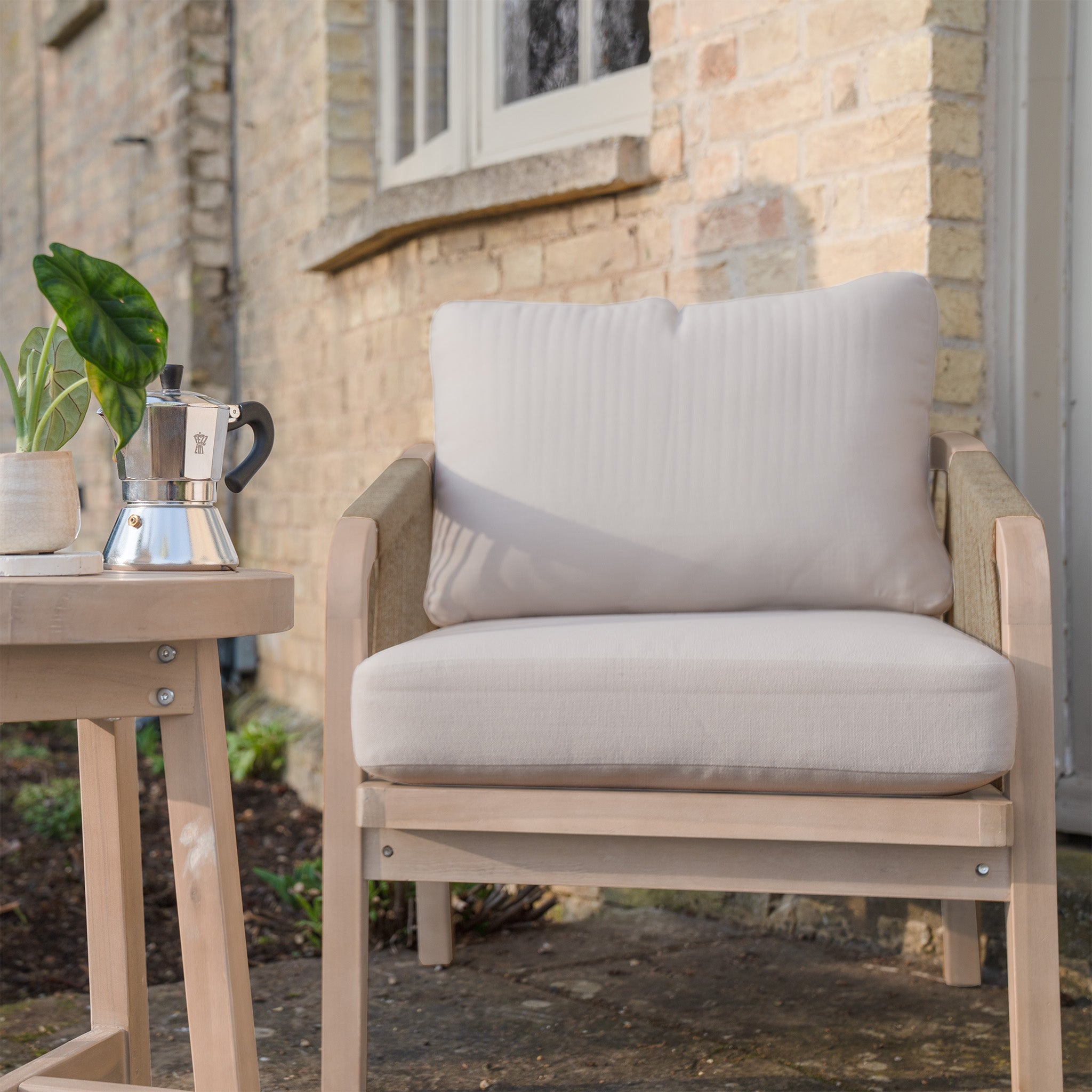 Mocha cushioned outdoor chair from the Quay Lounge Set sits beside a small table with a plant and coffee pot, placed by a brick wall—ideal garden furniture for relaxing outside.