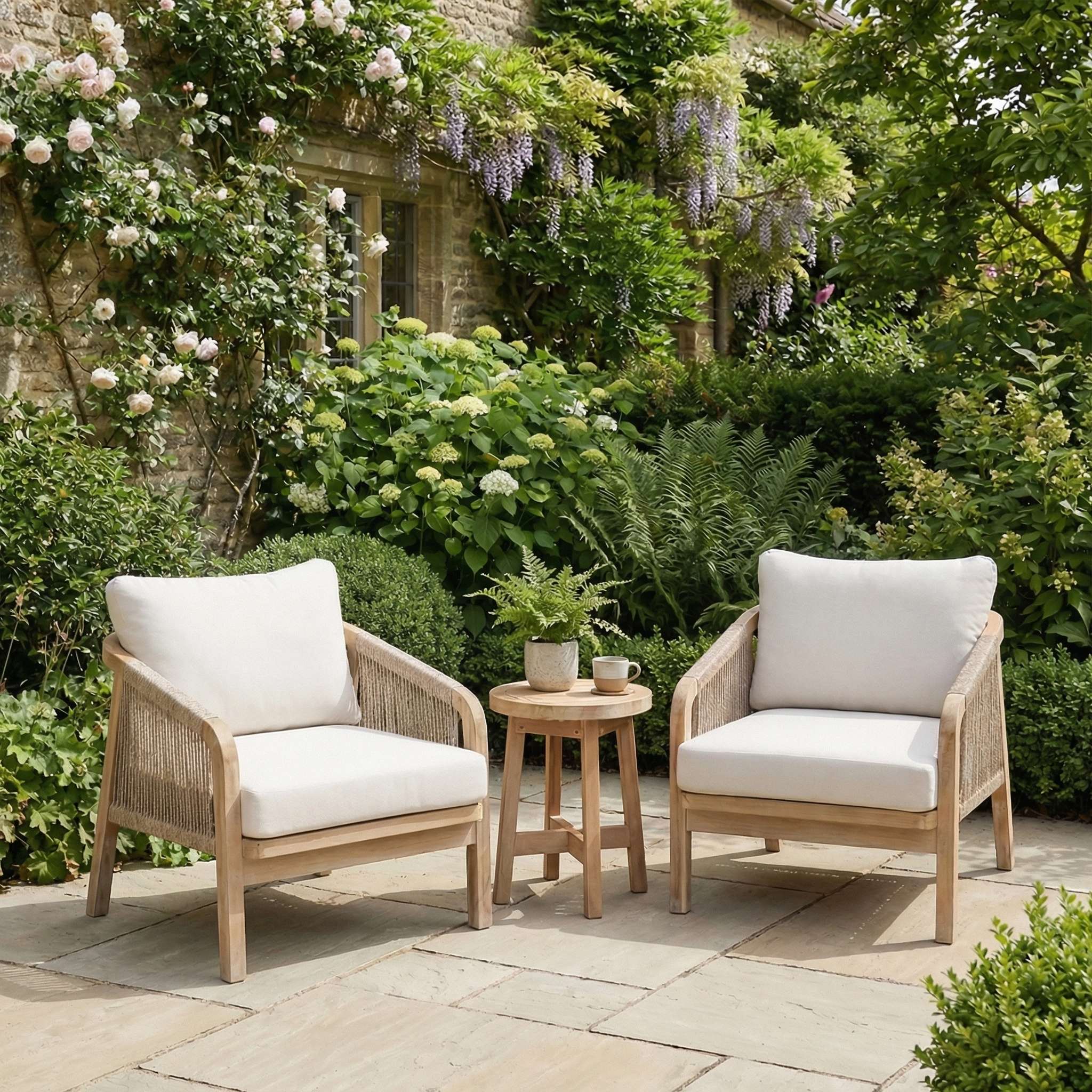 The Quay Lounge Set in Linen, featuring two cushioned acacia wood chairs with rope weave backs and a small table, creates an inviting outdoor lounging area surrounded by lush garden greenery and flowering plants.