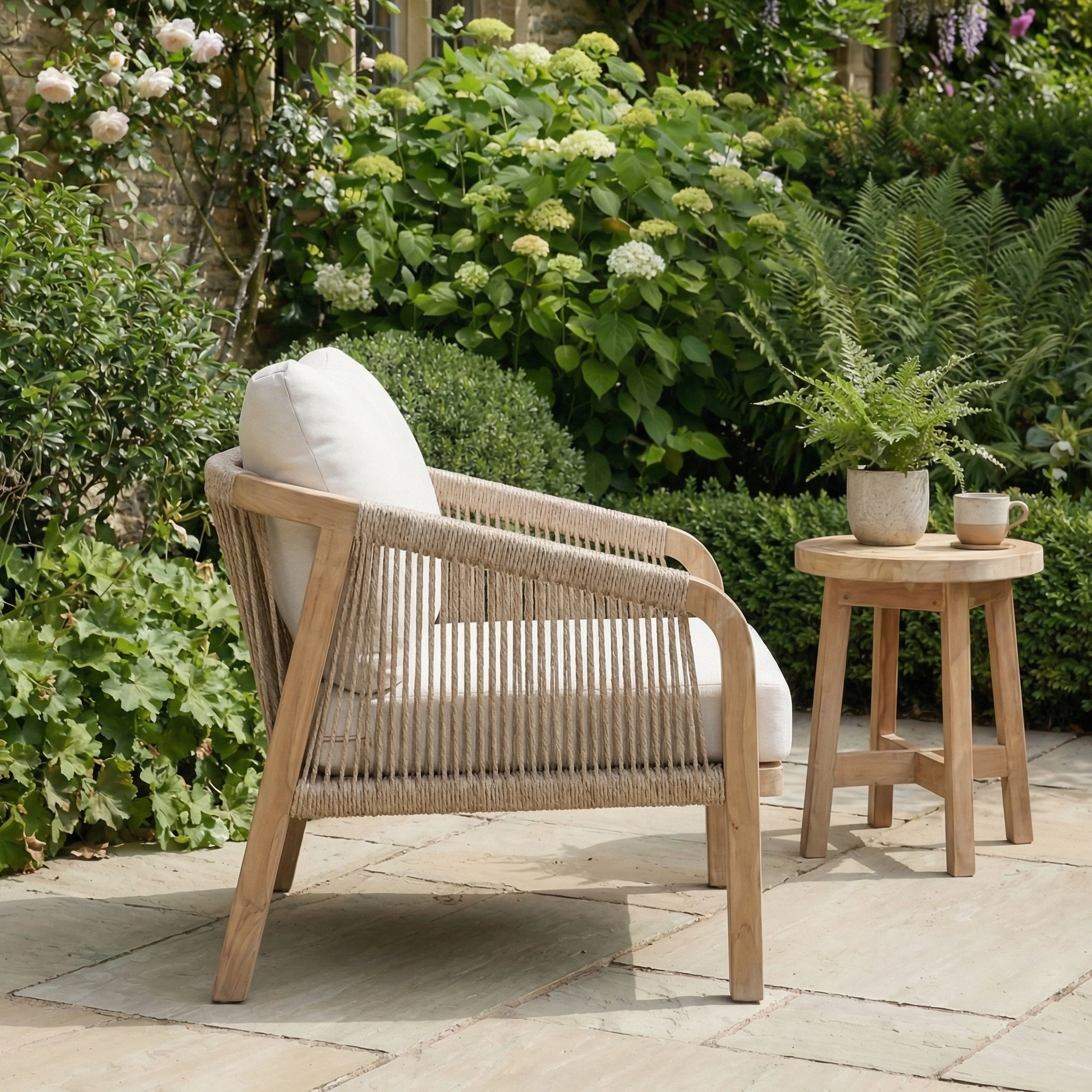 The Quay Lounge Set In Linen, featuring a chair with a rope weave back and white cushion plus a round side table, creates a cozy outdoor retreat on your stone patio amid lush greenery.