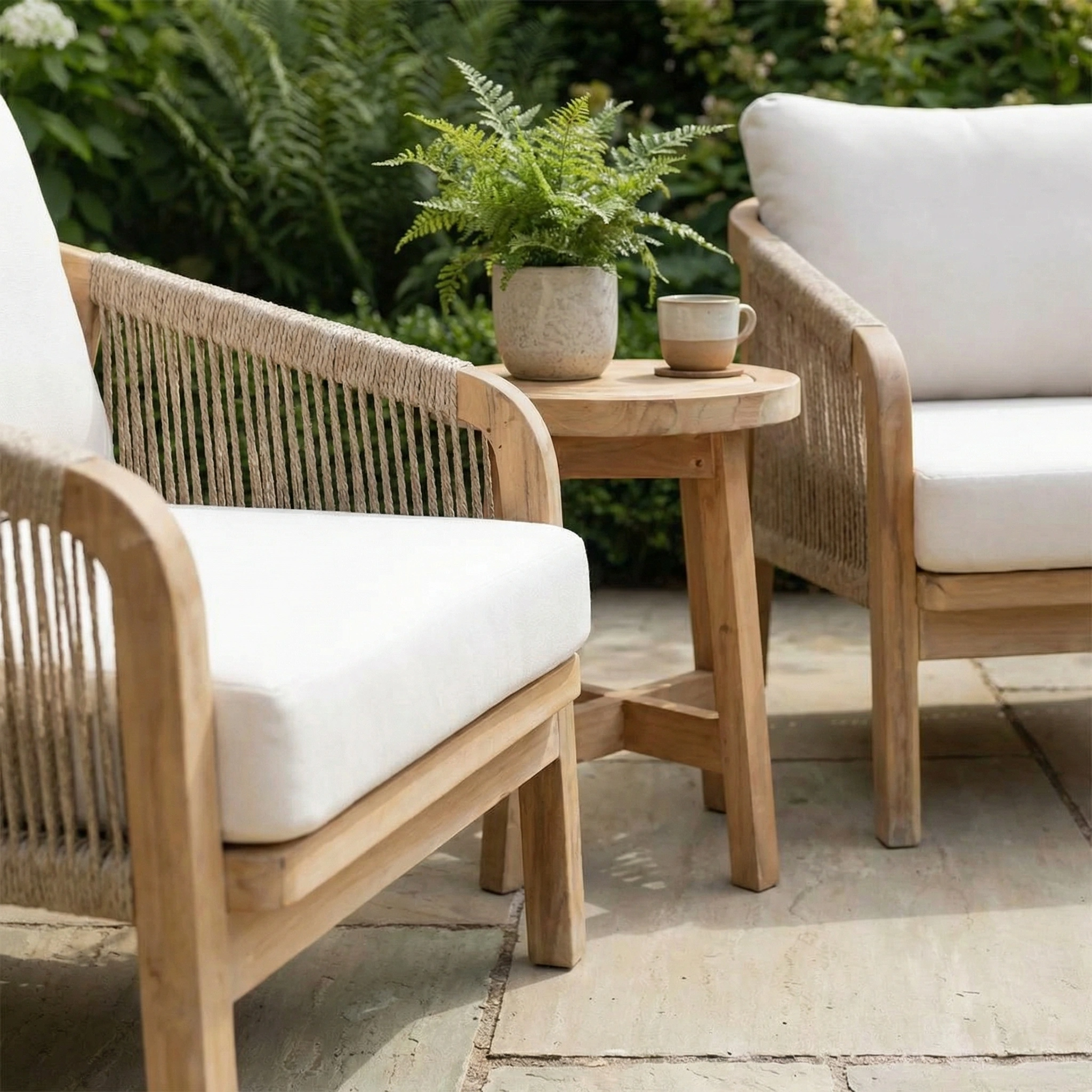 The Quay Lounge Set In Linen, featuring two wooden chairs with rope-weave backs, white cushions, and a small table, creates an inviting outdoor lounging area.