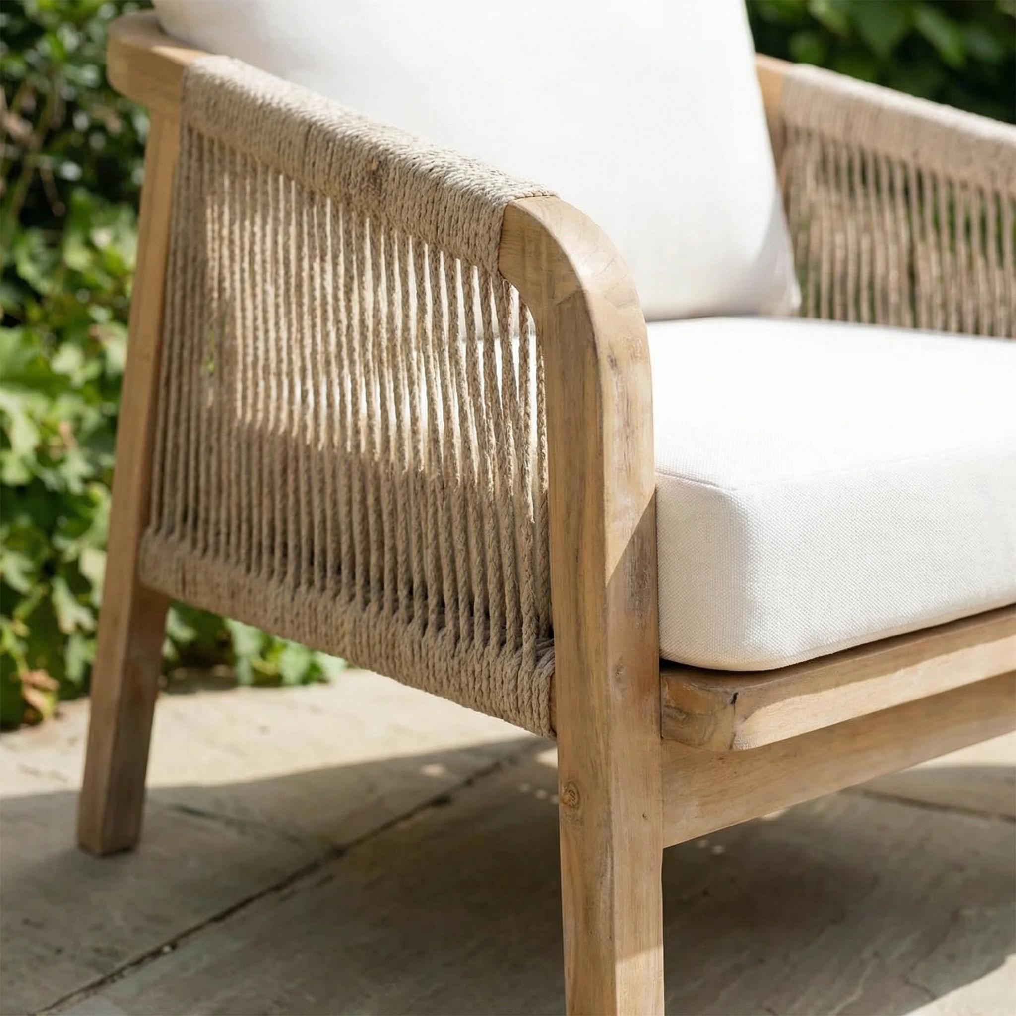 The Quay Lounge Set In Linen features a wooden outdoor chair with a rope-weave back and white cushions, made from acacia wood, styled on a stone patio with lush greenery creating an inviting lounging area.
