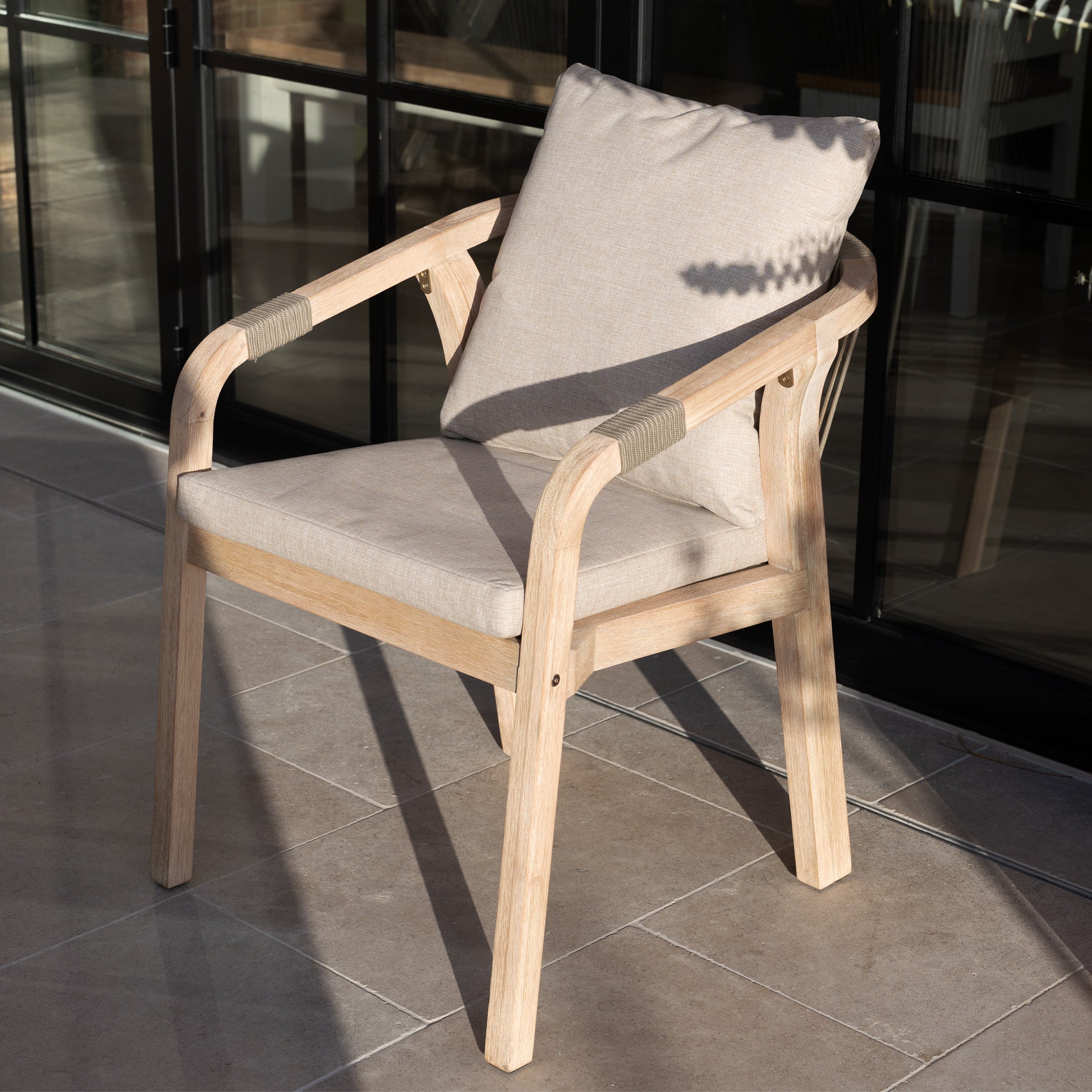 The Quay 4 Seat Round Dining Set in Mocha features cushioned wooden chairs and a pillow, perfect for relaxing on a tiled patio as sunlight casts elegant shadows across your outdoor space.