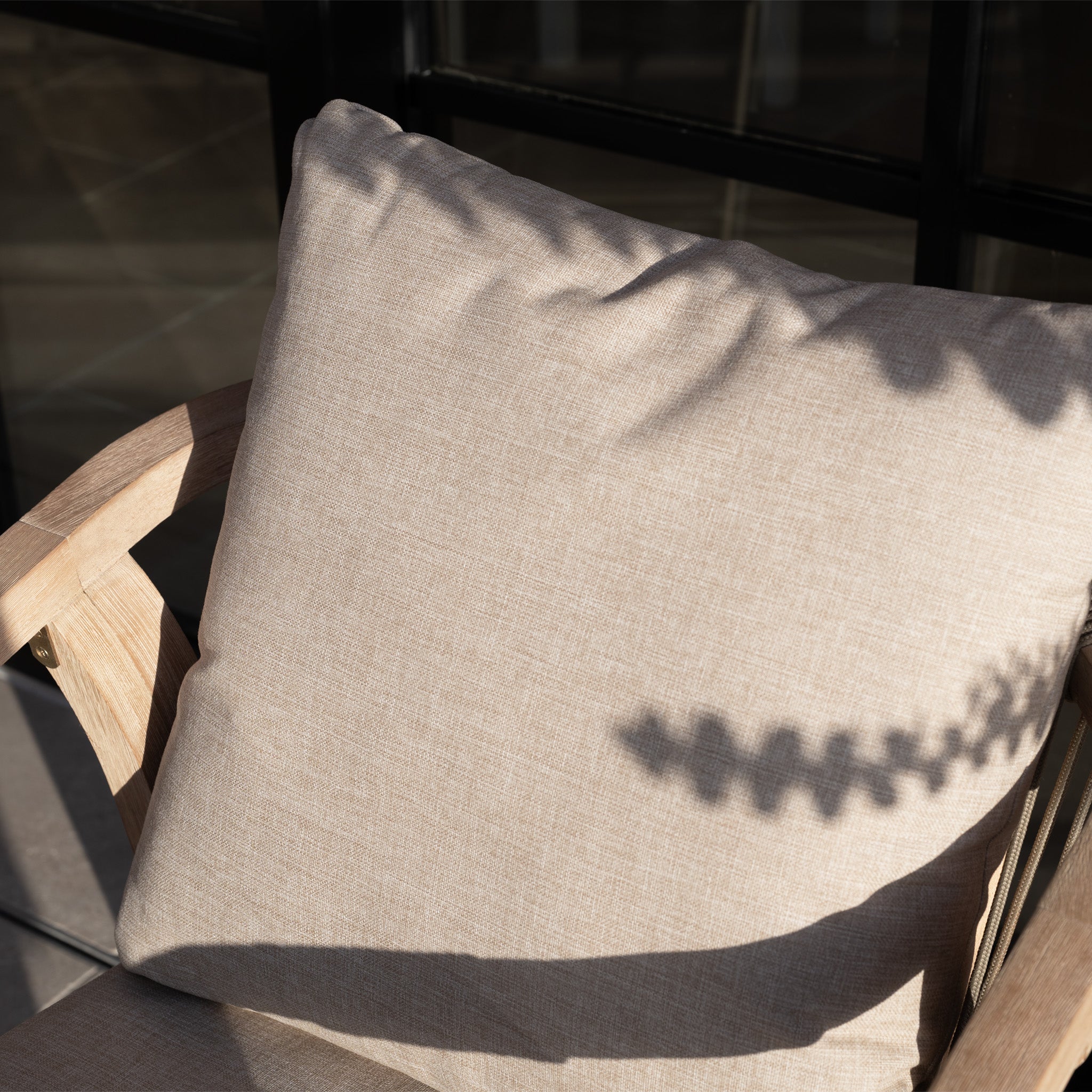 Beige cushion on a wooden chair from the Quay 4 Seat Round Dining Set in Mocha, with leafy shadows cast by sunlight.