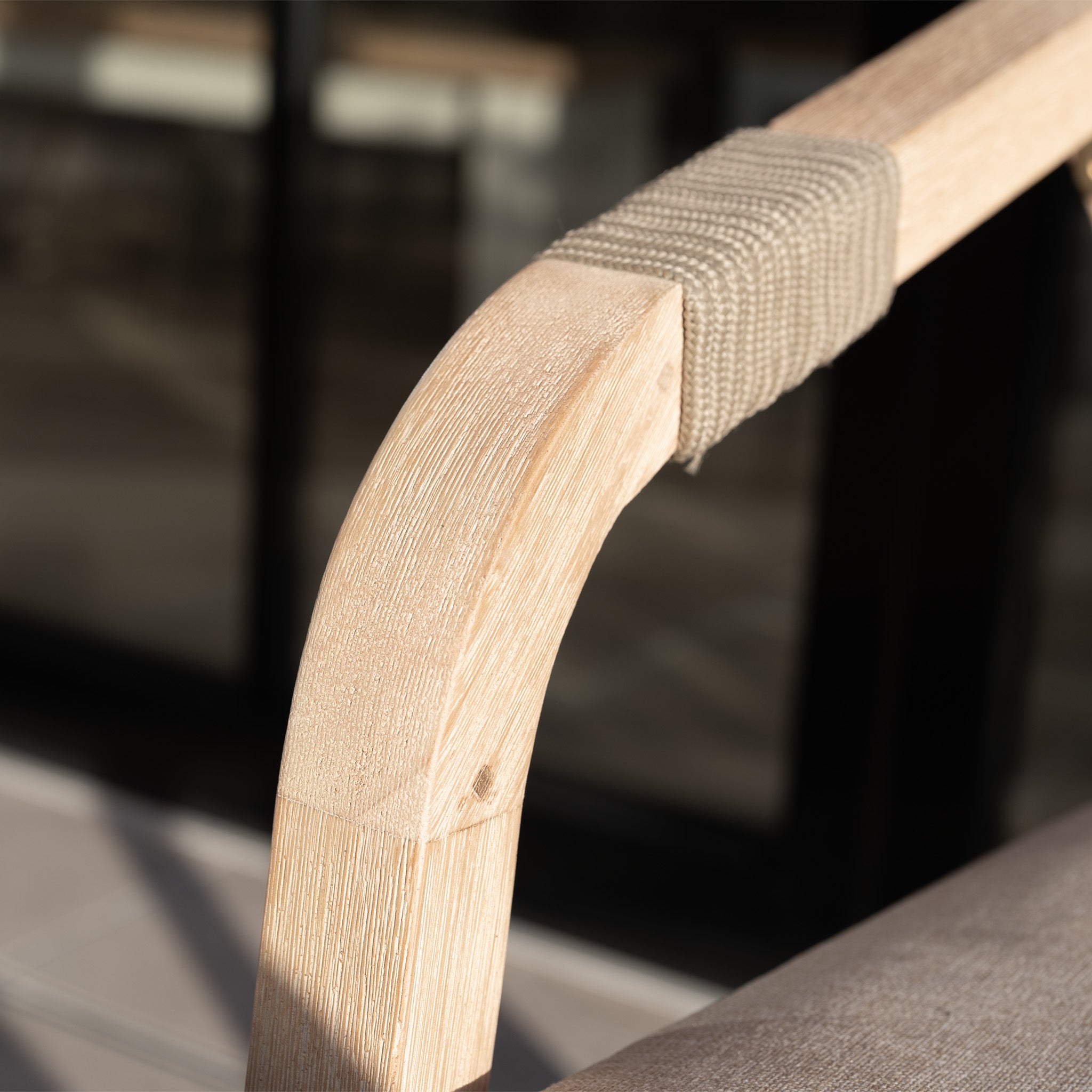 Close-up of a light wooden armrest with rope detail from the Quay 4 Seat Round Dining Set in Mocha, set against a blurred glass background.