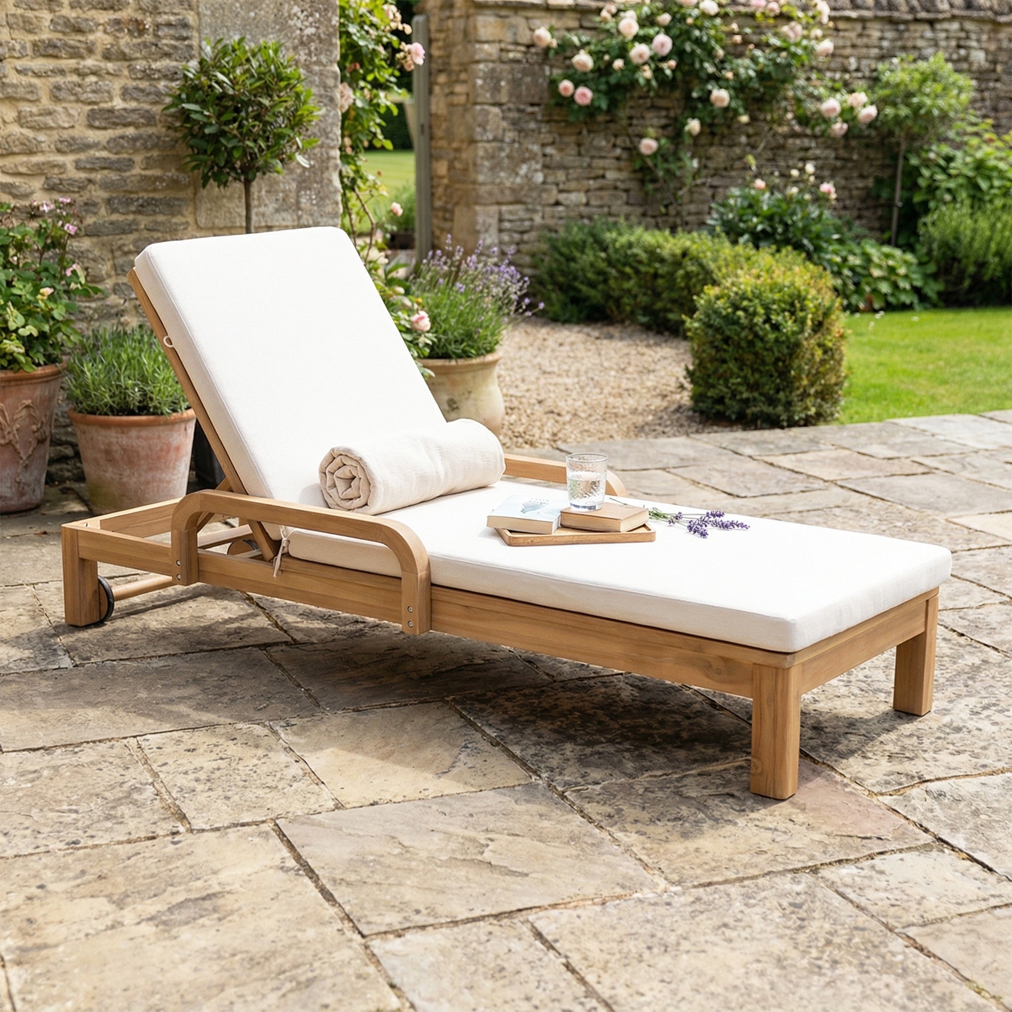 The Quay Sun Lounger in Linen features white cushions on acacia wood, set on a stone patio with potted plants and garden views—perfect outdoor furniture for relaxing.