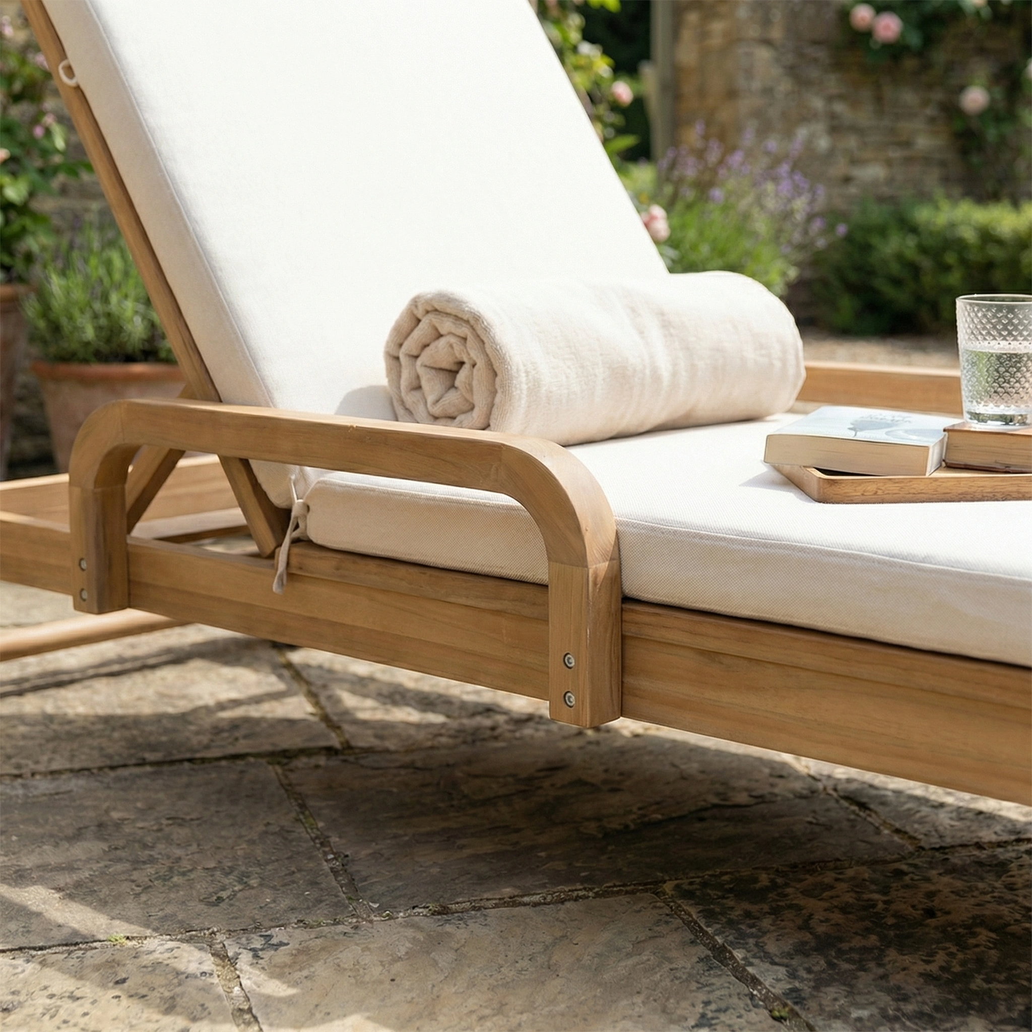The Quay Sun Lounger in Linen sits invitingly on a stone patio with a rolled towel, glass of water, and book—outdoor furniture perfect for relaxation.