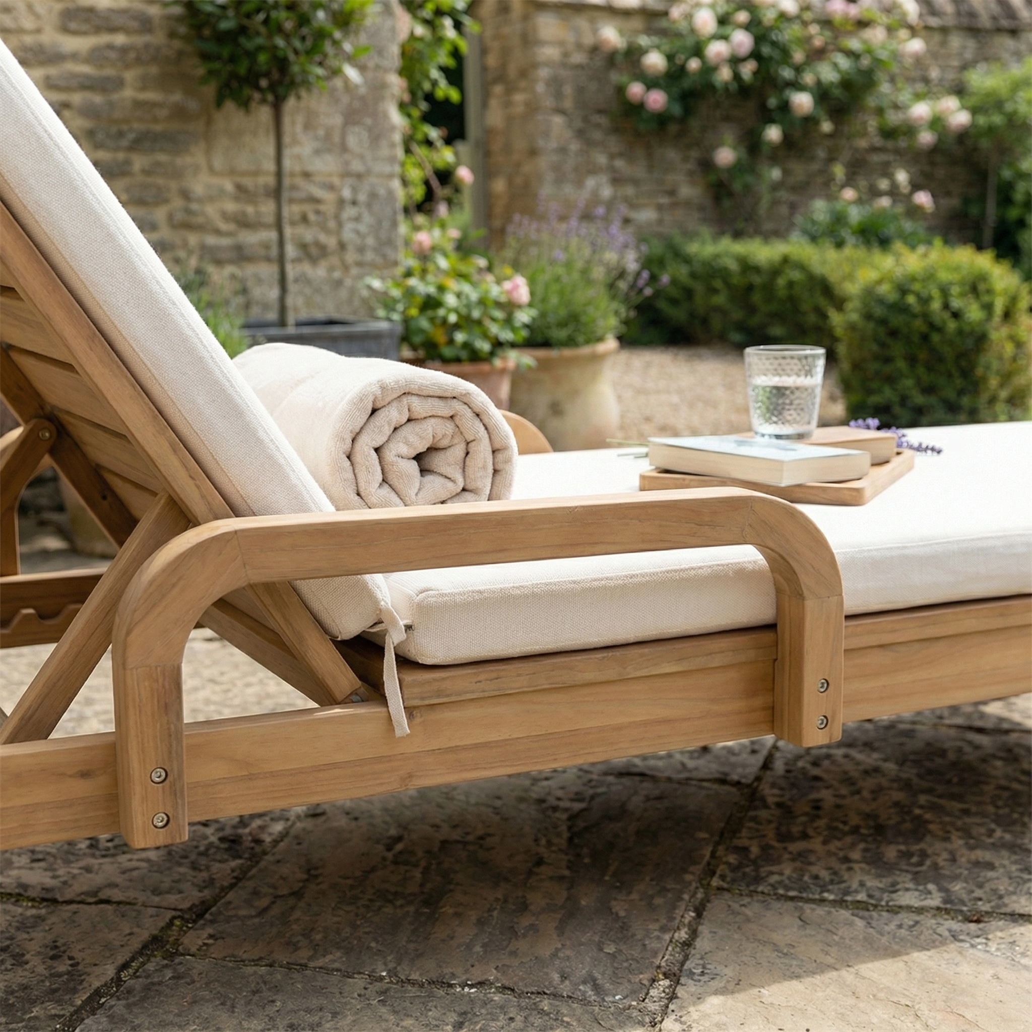The Quay Sun Lounger in Linen awaits on a stone patio, complete with cushion, rolled towel, books, and a glass of water—perfect for relaxing in your garden.