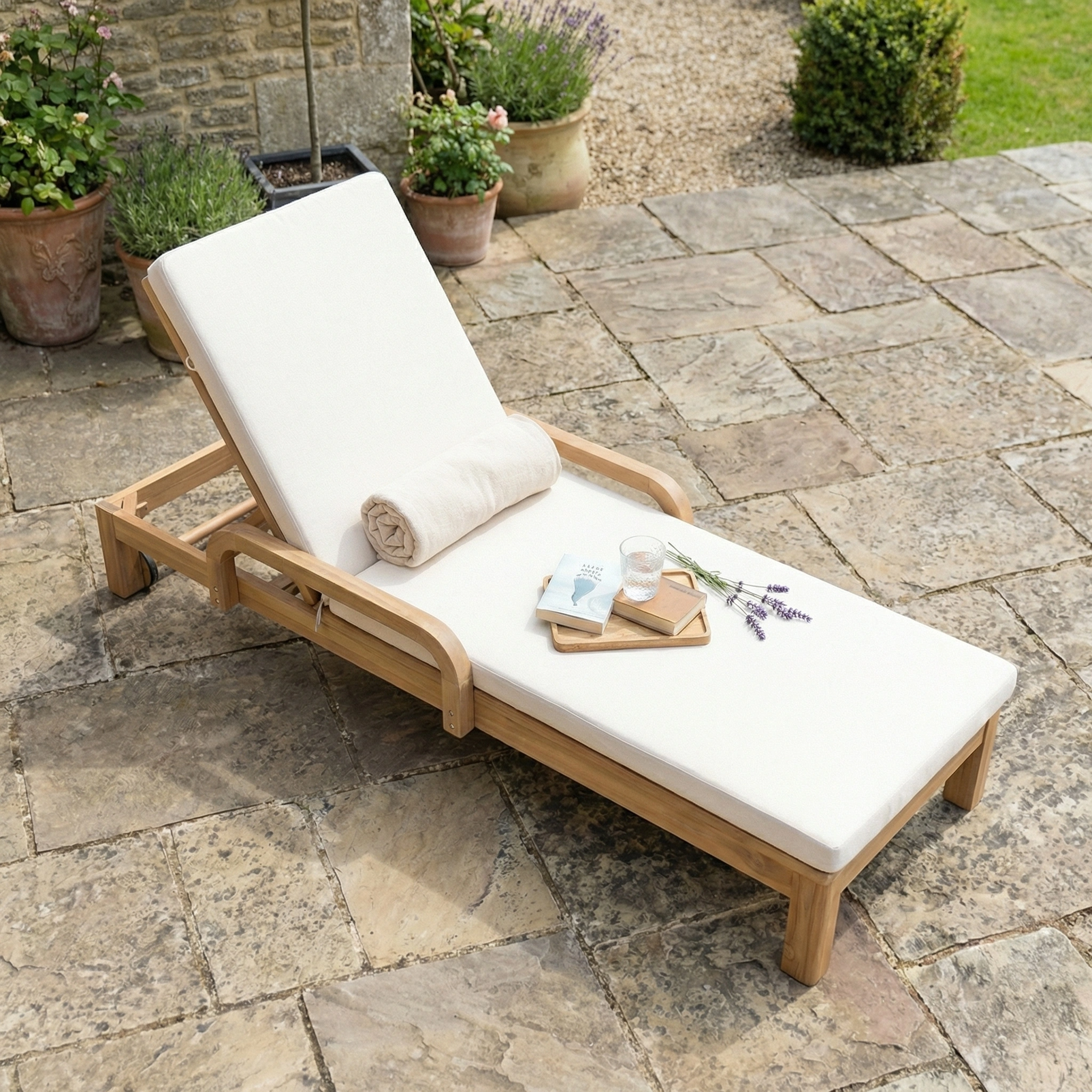 Quay Sun Lounger In Linen features white cushions, a rolled towel, and a tray with a drink, book, and lavender on a stone patio—stylish outdoor furniture for relaxing in comfort.