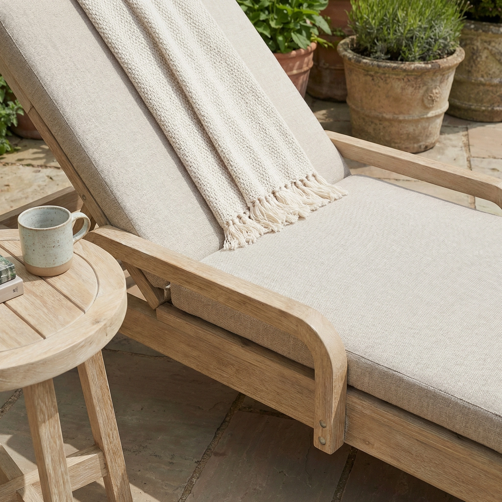 Mocha cushioned Quay Sun Lounger with a throw blanket beside a small round table and potted plants, perfect as stylish garden furniture for outdoor relaxation.