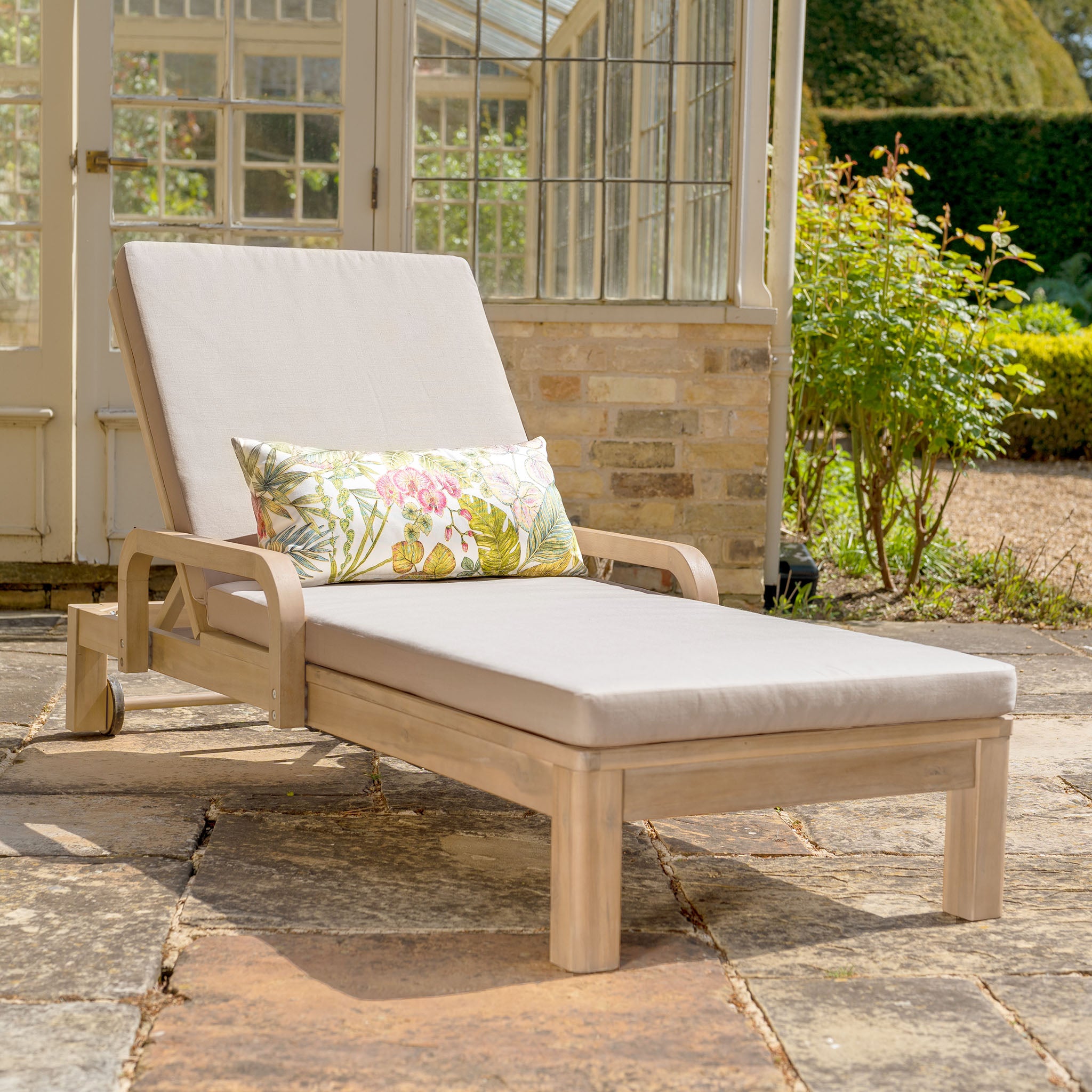 The Quay Sun Lounger in Mocha features a cushioned seat and sits on a stone patio near glass doors and lush garden plants—perfect outdoor furniture for stylish relaxation.
