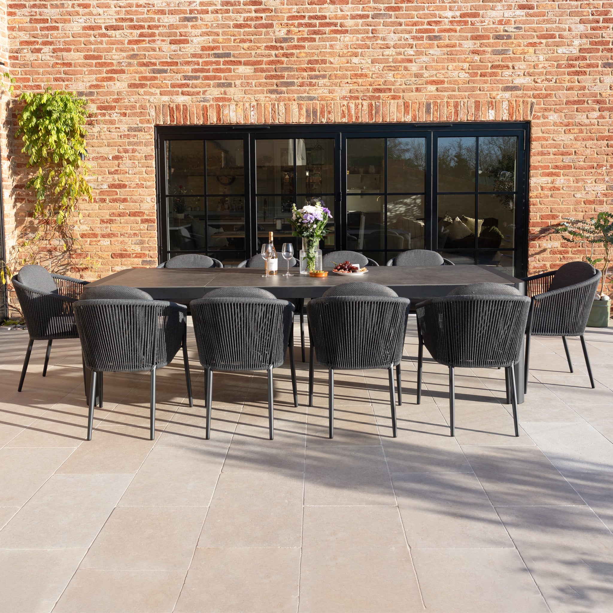 The Ravona 10 Seat Rope Extending Dining Set (207-277cm) in Charcoal is arranged for a meal, adding an elegant touch to a patio beside a brick wall and glass doors.