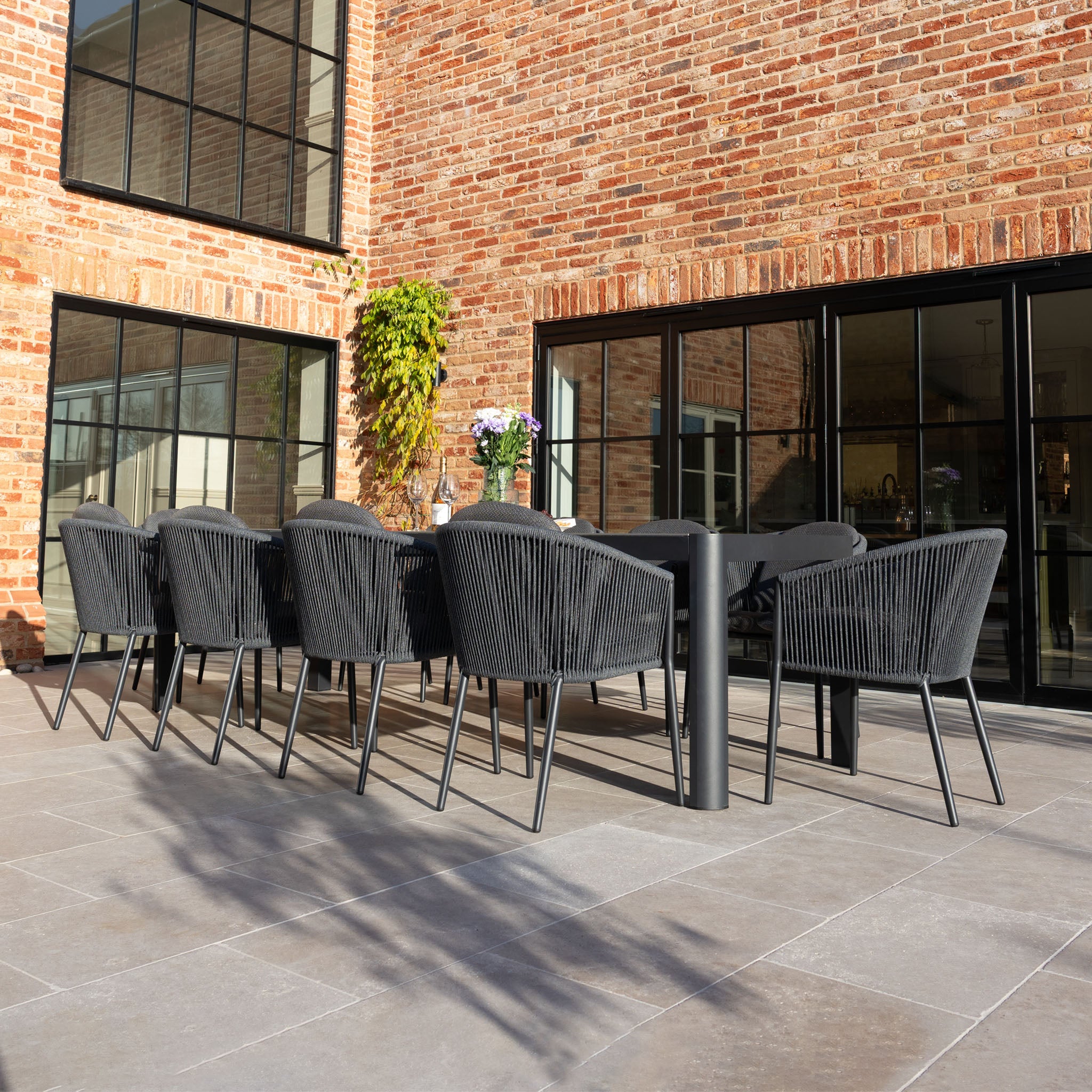 Modern outdoor patio with a Ravona 10 Seat Rope Extending Dining Set (207-277cm) in Charcoal, featuring a black dining table, ten chairs, and brick walls with large windows.