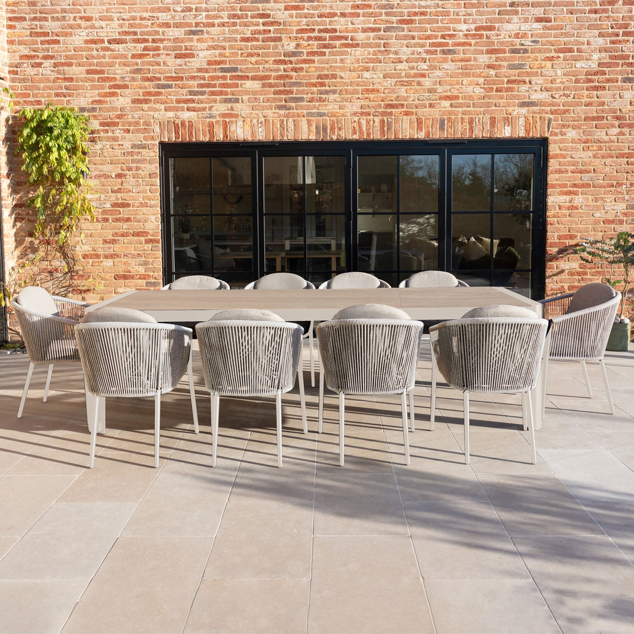 The Ravona 10 Seat Rope Extending Dining Set (207-277cm) in Latte, shown on a tiled patio by glass doors and a brick wall, is perfect for stylish outdoor gatherings.