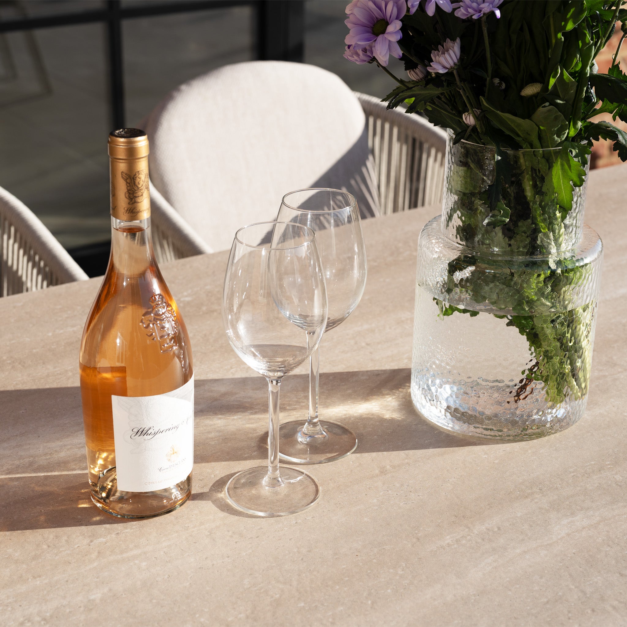 A bottle of rosé, two glasses, and a vase of flowers sit on a sunlit table with an elegant aluminium frame—perfect for gatherings around the Ravona 6 Seat Rope Extending Dining Set (162-222cm) in Latte.