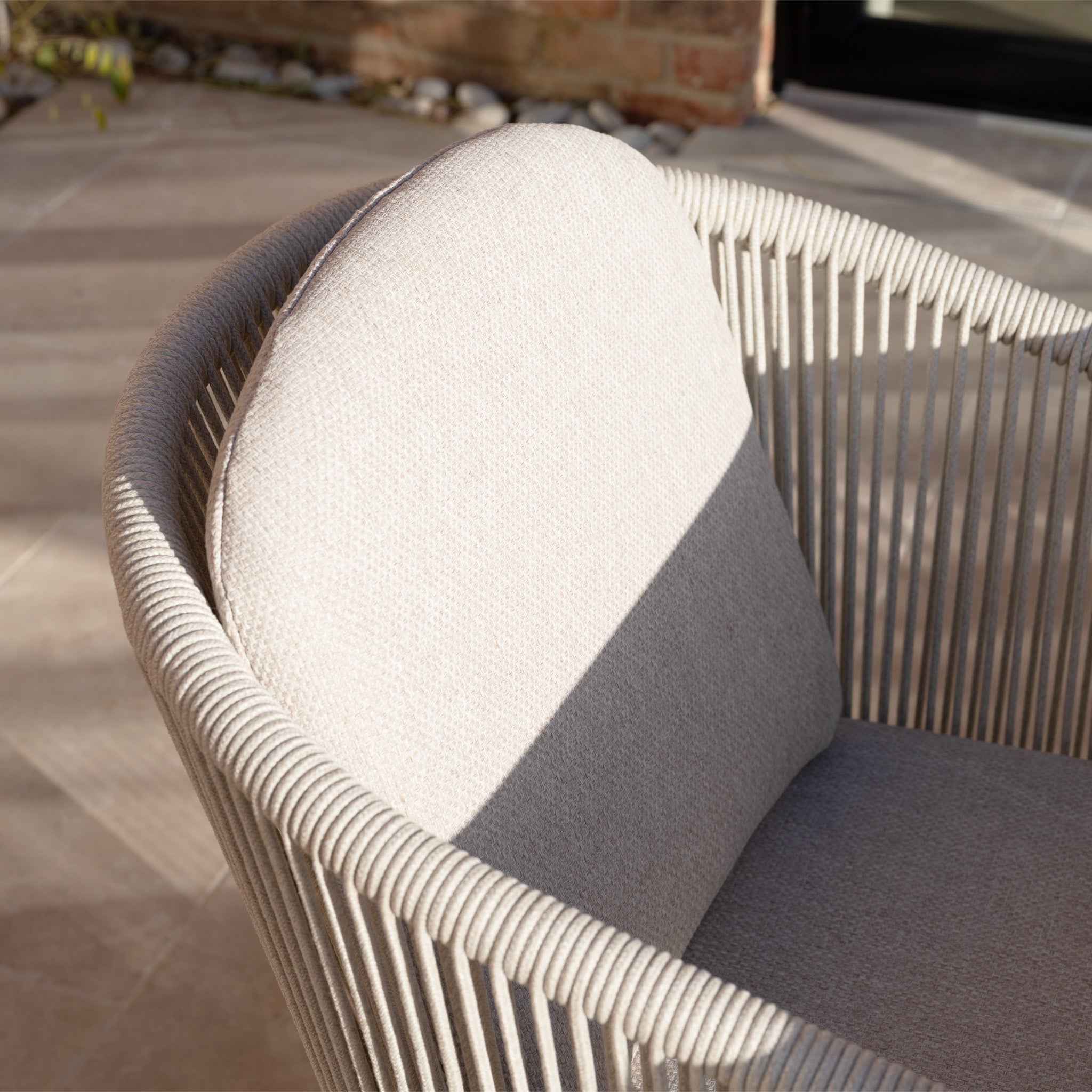 Close-up of a light beige woven chair from the Ravona 6 Seat Rope Extending Dining Set (162-222cm) in Latte, showing its aluminium frame highlighted by sunlight and shadow on the seat and backrest.
