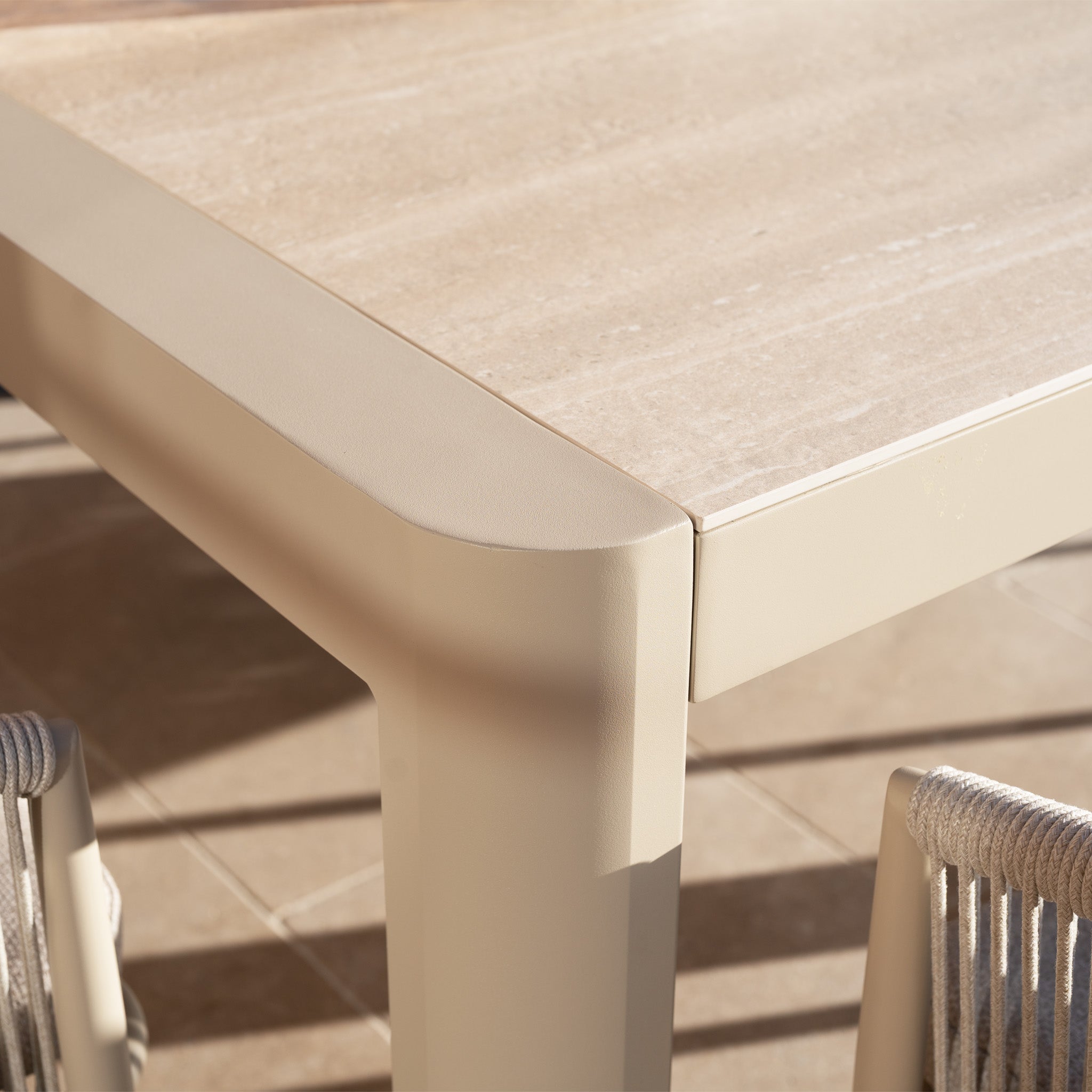 The Ravona 6 Seat Rope Extending Dining Set (162-222cm) in Latte features a smooth stone top table and rope-woven chairs with aluminium frames, offering stylish outdoor dining on a sunlit tiled patio.