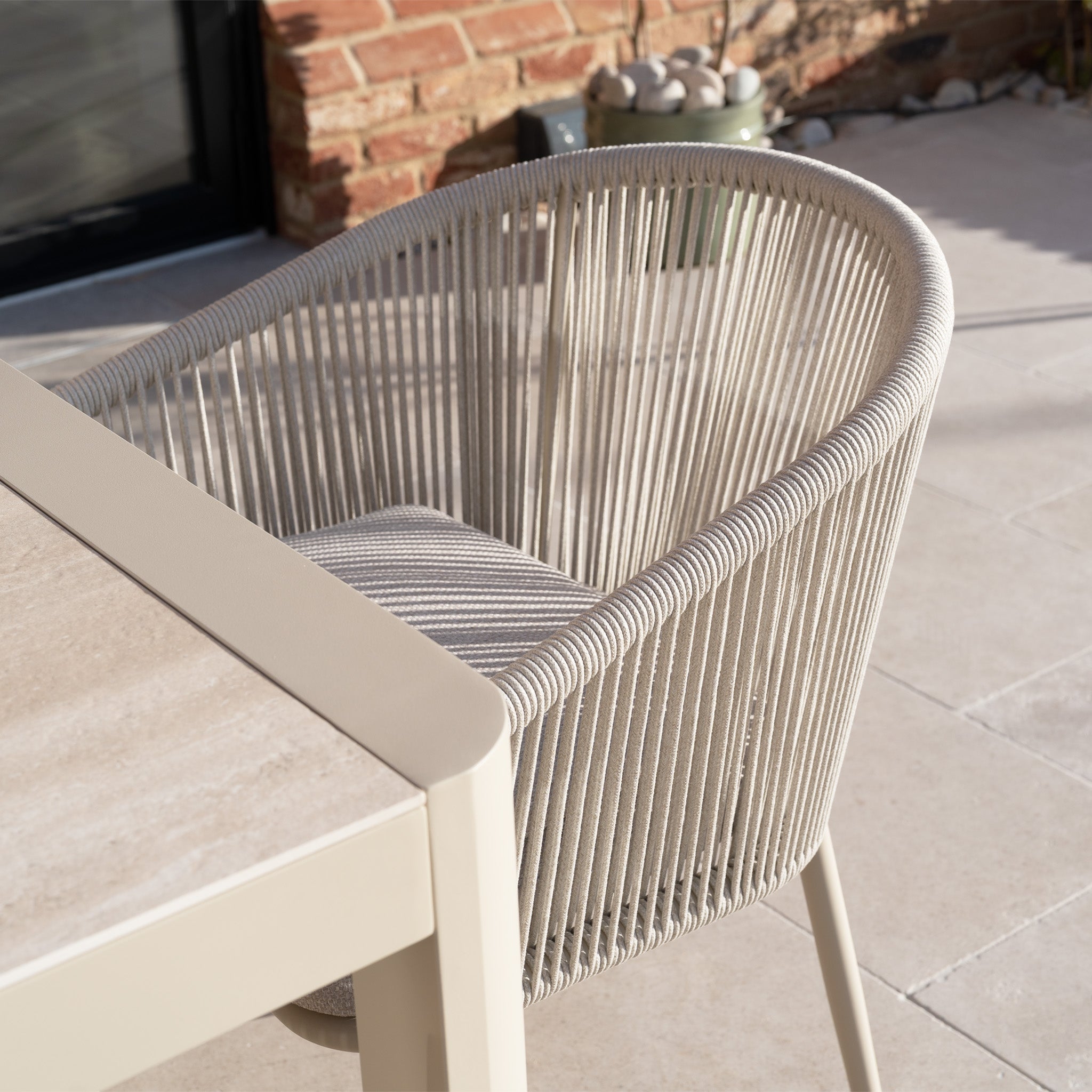 A beige rope outdoor chair with an aluminium frame sits by a light table, both part of the Ravona 6 Seat Rope Extending Dining Set (162-222cm) in Latte, on a sunlit tiled patio.