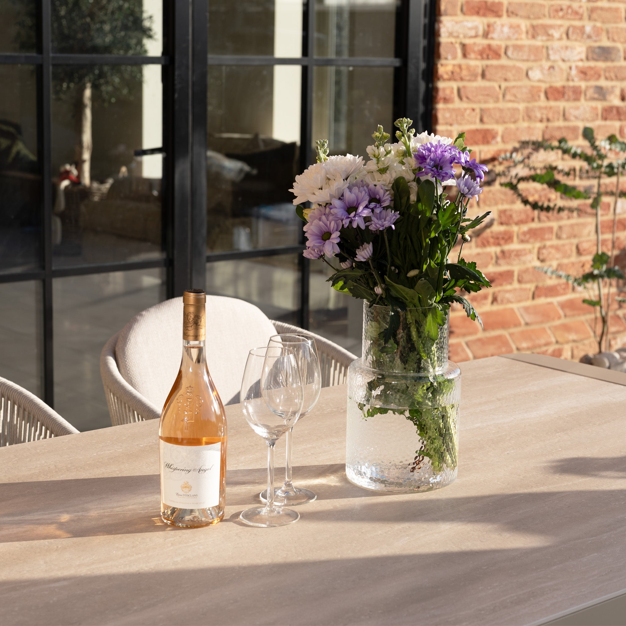 A bottle of rosé, two glasses, and a vase of purple and white flowers rest on the Ravona 6 Seat Rope Extending Dining Set (162-222cm) in Latte, creating an inviting scene atop its sleek aluminium outdoor table.